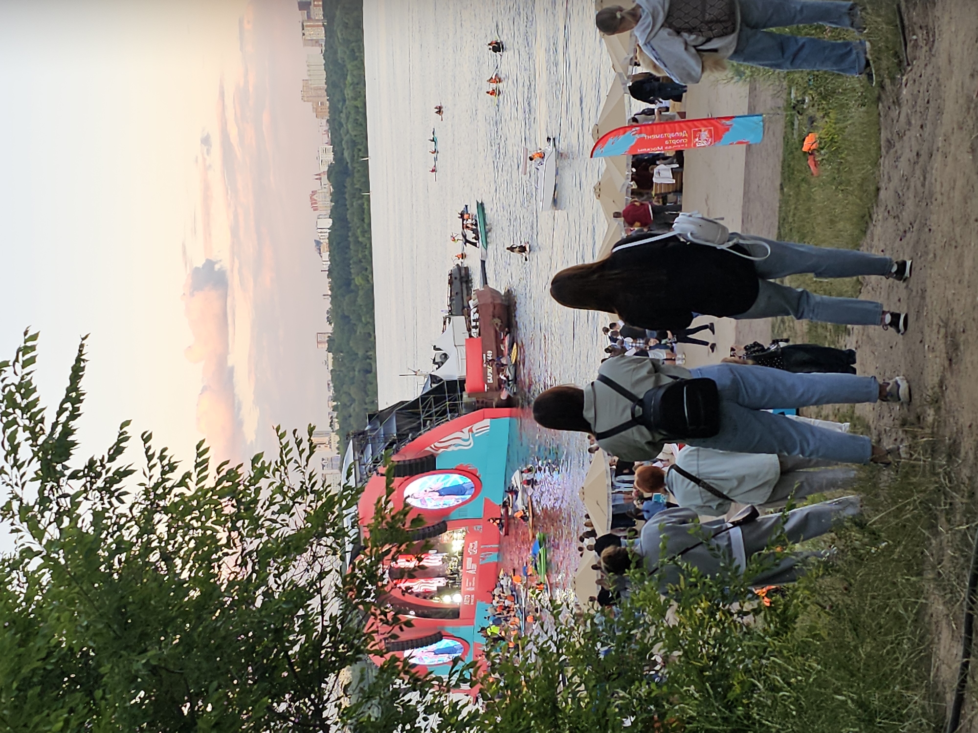 This image depicts an outdoor event on a riverbank in Khimki, Russia, likely during the evening, as suggested by the soft, pastel hues of the sky. In the foreground, a group of people are gathered, viewed from behind, facing a large, elaborate stage set up on the water. The stage features a curved, red and teal structure with multiple screens displaying images of performers. The river is populated with numerous individuals on inflatable rafts, kayaks, and paddleboards, some of whom are in close proximity to the stage.
Further out on the water, a variety of boats are visible, including a white speedboat with an orange-clad occupant and several darker vessels. The riverbank behind the stage is lined with beach umbrellas and a crowd of spectators. A dense line of trees forms a backdrop against the city skyline, which features several buildings. Lush green foliage from trees in the immediate foreground partially obscures the view. The overall atmosphere suggests a lively gathering with a focus on water-based activities and a performance on a unique floating stage.