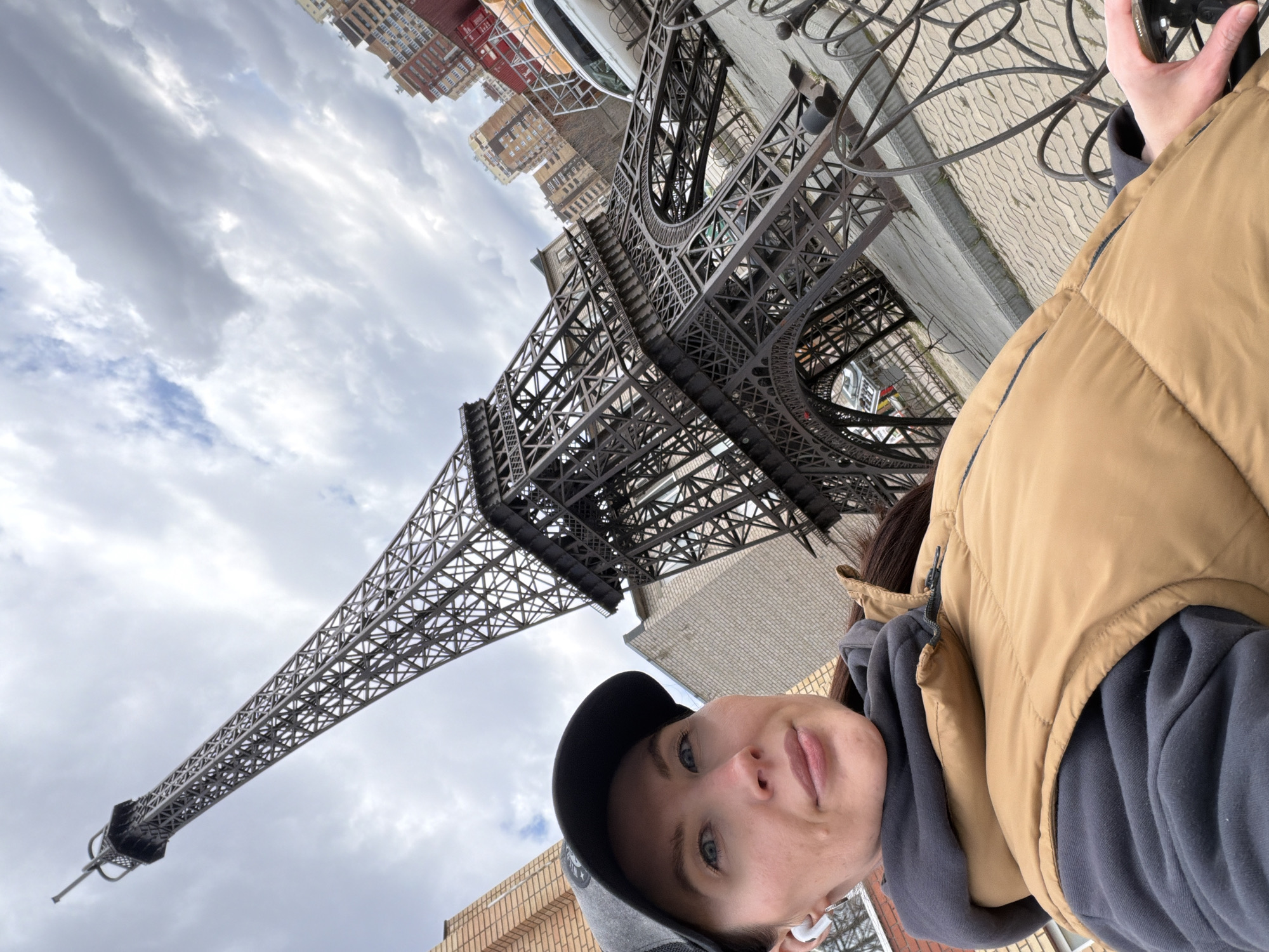 A woman is captured in a selfie-style shot, looking directly at the camera, with a prominent replica of the Eiffel Tower behind her. The image is taken from a low angle, looking upwards towards the sky and the top of the tower.
The subject is a woman, visible from the chest up. She wears a black baseball cap, a dark grey hooded sweatshirt, and a tan puffer vest. Her light-colored eyes are visible, and her expression is neutral or slightly pleasant. White wireless earbuds are noticeable in her ears. Her right hand is partially visible in the upper right corner, appearing to hold the camera.
The central feature in the background is a large, detailed replica of the Eiffel Tower, made of dark metal. Its intricate lattice structure is clearly visible, suggesting it's a significant public art piece or landmark. The tower extends upwards, partially obscured by the woman, reaching high into the cloudy sky.
The sky is overcast with large, grey clouds, indicating a daytime scene. In the distance, behind the tower, several multi-story brick buildings, varying in color, line an urban street. To the right of the woman's head, a paved area with a decorative pattern, possibly cobblestones, and a section of an ornate metal fence are visible.
The scene depicts a casual moment, likely a tourist or local, posing with a well-known architectural reproduction in an urban environment, specifically identified as Perm, Russia. No visible text is present in the image.