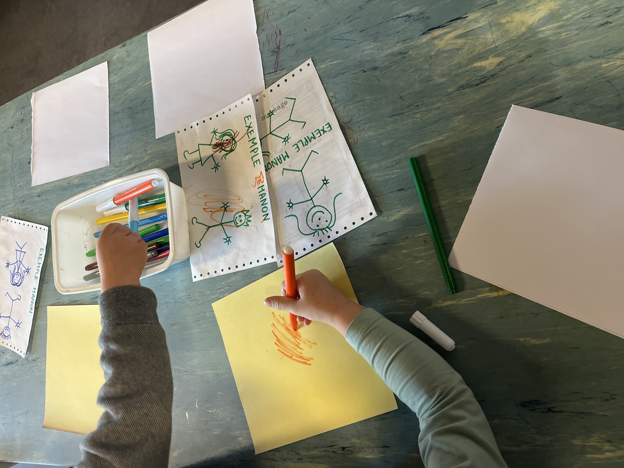 The scene is a top-down view of a child drawing on a yellow piece of paper with an orange marker. The child is wearing a gray shirt with their hands visible. On the table are other pieces of paper, some with stick figure drawings and the words "EXEMPLE MANON" (Example Manon) written in French. There is a white container filled with markers, a green pencil, and several blank pieces of paper. The table has a green and blue marble-like surface. The location is likely a residence or school in La Fouillouse, France based on the French writing.