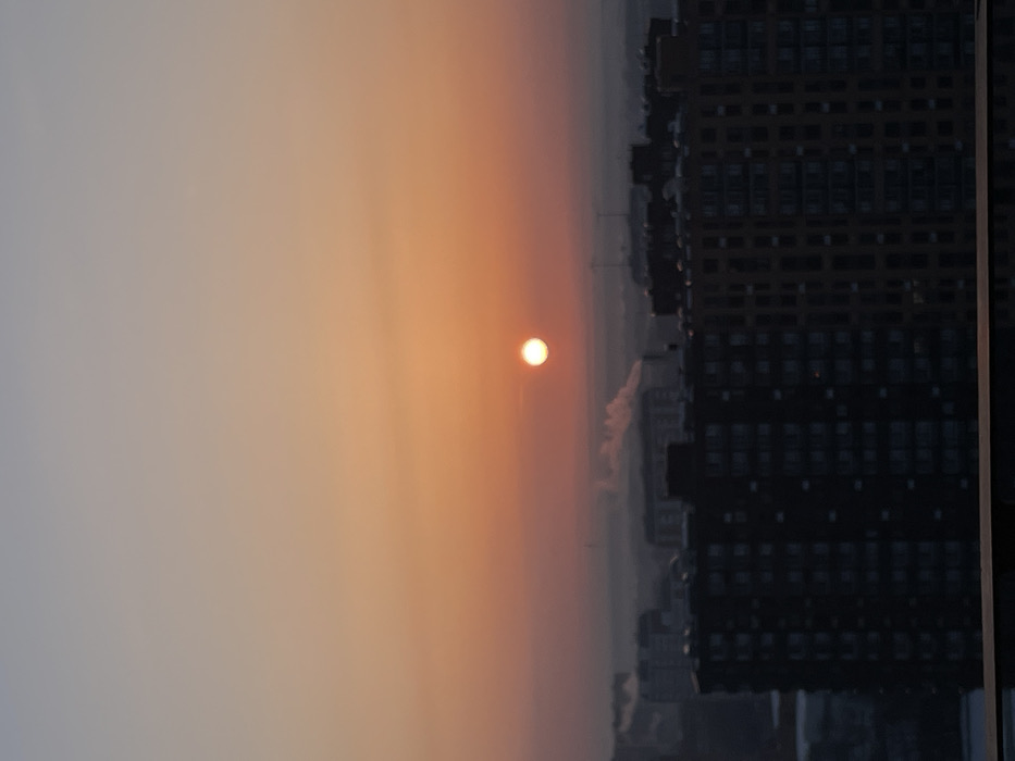 This image depicts an elevated panoramic view over the city of Elektrougli, Russia, at either sunrise or sunset. The sky transitions from a pale blue-gray at the top to vibrant orange and red hues towards the horizon. A prominent, bright, circular, orange-white sun is situated in the central sky area. The immediate foreground features the dark silhouettes of multiple multi-story residential buildings, some dark gray-black and others a muted brown-orange, obscuring the lower portions of the distant cityscape. A widespread atmospheric haze or fog blankets the mid-distance urban landscape. Within this hazy layer, distinct white plumes of smoke are observed rising from at least two separate, indistinct sources. No individuals, specific actions, or interactions are discernible. The environment is an urban setting characterized by densely packed residential structures and visible industrial or heating emissions.