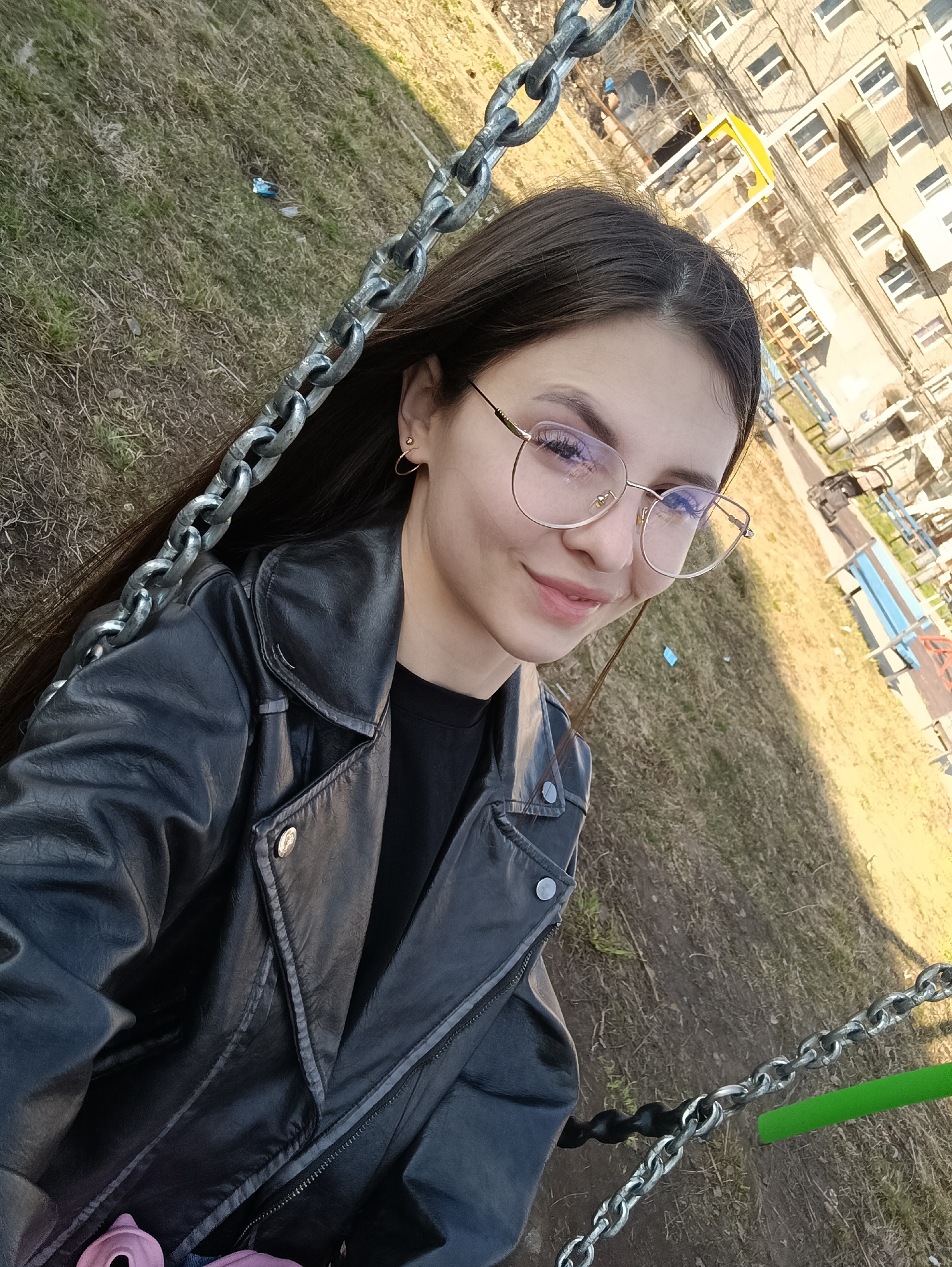 A young woman is taking a selfie while on a swing set outdoors. The background shows a multi-story apartment building with beige walls and multiple windows. The foreground is dominated by the woman, who has long, dark hair and is wearing gold-rimmed glasses and a black leather jacket over a black shirt. The chain of the swing set is visible on both sides of the woman, made of metal and appearing heavily used. The ground surrounding the swing set consists of grassy areas with bare patches. In the background, there is also a playground area visible with benches. The location appears to be in an urban area in the city of Unknown, Unknown.