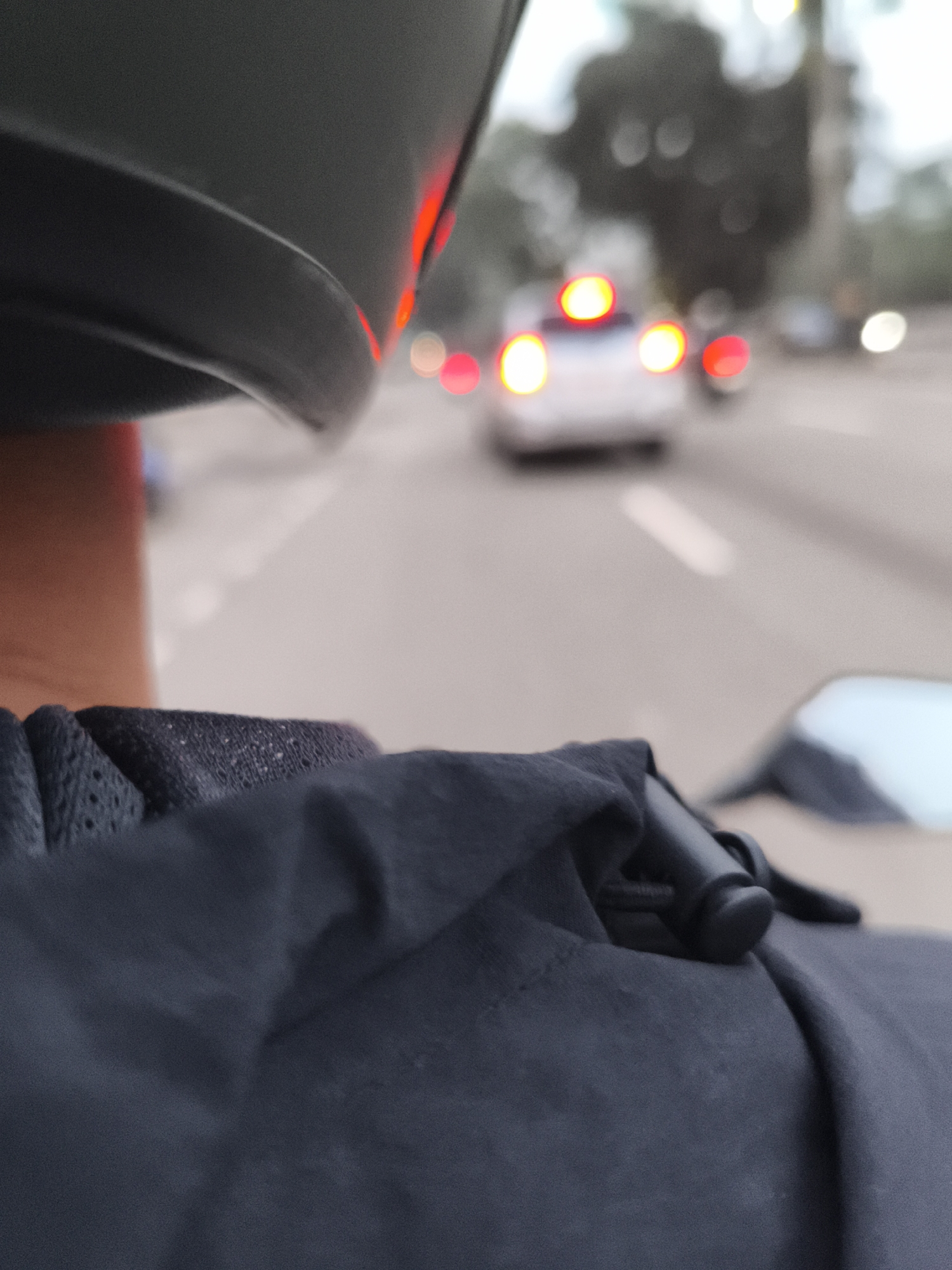 The image captures a first-person perspective from the back of a motorcycle, looking over the shoulder of the rider or a passenger. In the foreground, the top part of a dark grey helmet fills the upper left corner, while the bottom half of the frame is dominated by dark grey or black fabric, likely a jacket or backpack, featuring a visible black drawstring toggle on the right. A section of reddish-brown skin, possibly a neck or shoulder, is visible between the helmet and the garment.
The scene ahead depicts a road with several vehicles, all rendered in a soft blur due to shallow depth of field. The most distinct elements in the mid-ground are the bright red and amber taillights of cars, particularly a white car centrally positioned ahead with its lights illuminated. The road itself is grey with subtle white lane markings. The background consists of indistinct, blurry shapes of trees or foliage against a muted, light grey sky.
The prominent glowing taillights strongly suggest that the activity of riding the motorcycle is taking place during dusk, dawn, or nighttime, or possibly under very overcast conditions. The setting appears to be a road within a semi-urban or suburban area, consistent with the provided location context of Sukaraja, Indonesia. There is no visible text in the image.