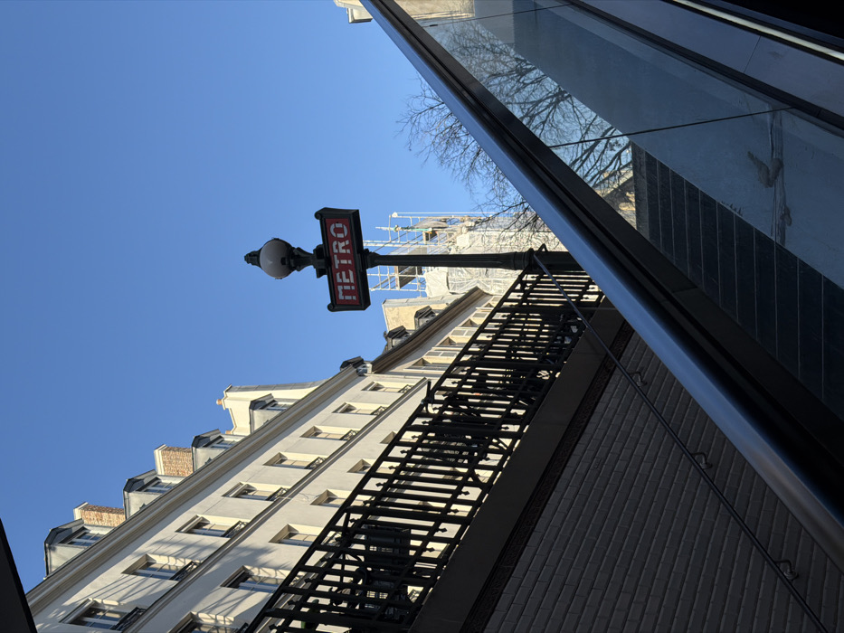 The scene depicts an upward perspective from the entrance of a Paris Métro station under a clear, bright blue sky. A traditional black lamppost with an illuminated red and white "METRO" sign is centrally located, signifying the station. To the right, a modern escalator with a silver metal handrail and transparent glass balustrades ascends, paralleled by a set of dark stairs. On the left, a multi-story cream-colored building with numerous windows and gray mansard roofs is visible, fronted by a dark, ornate metal railing atop a grey brick wall. In the mid-ground, behind the Métro sign, a building under construction is partially visible, encased in white scaffolding. Bare tree branches are present on the right. No individuals are depicted, and no specific actions or interactions are occurring. The environment is urban and well-lit by direct sunlight.