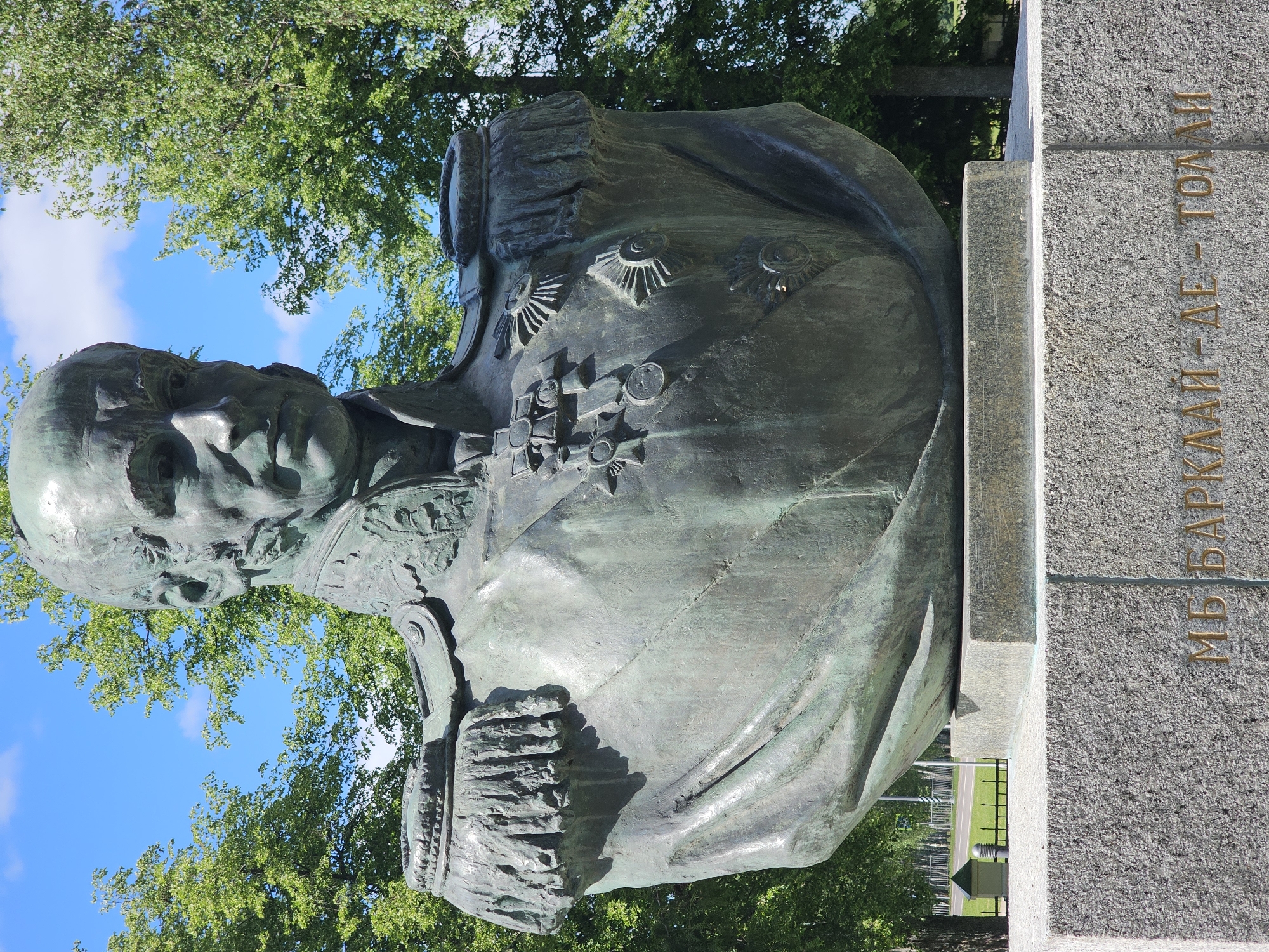 The image shows a bronze bust of Mikhail Bogdanovich Barclay de Tolly, a Russian Field Marshal, situated in Borodino, Russia. The bust depicts him in military attire adorned with medals and decorations. The monument is set against a backdrop of green trees and a blue sky with clouds. The base of the bust has the name "М.Б. БАРКЛАЙ-ДЕ-ТОЛЛИ" inscribed on it.