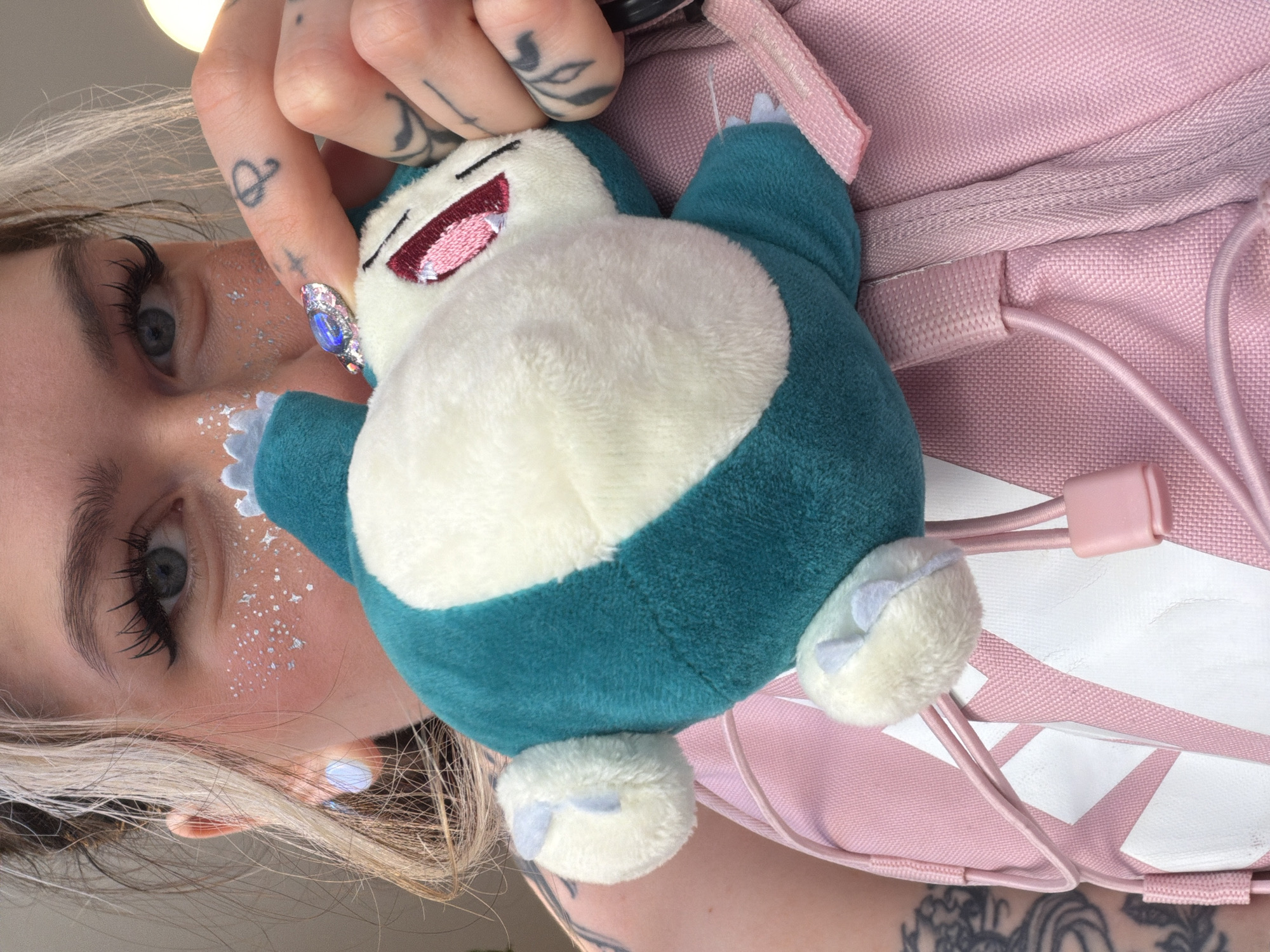 A young woman is holding a Snorlax plush toy. The woman has blond hair, and gray eyes, and her face is decorated with silver glitter. She has tattoos on her fingers and arm. The Snorlax is blue and white and has a smiling expression. The woman is wearing a pink backpack with a white stripe. The background is blurry and does not show any part of the city of Budva, Montenegro.