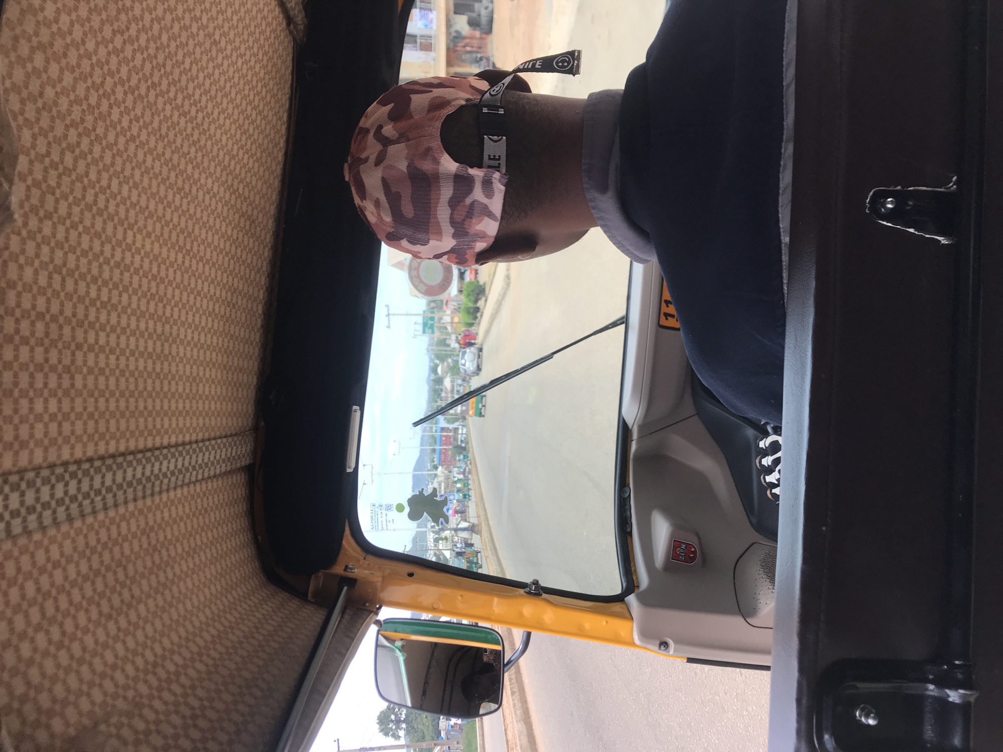The image depicts the interior and forward view from a yellow three-wheeled vehicle, likely an auto-rickshaw or "Keke Napep," in Rafin Par, Nigeria. The perspective is from the rear passenger compartment, looking over the driver's right shoulder through the windshield onto a road scene.
The vehicle's interior ceiling is upholstered in a light tan fabric with a repeating small checkerboard pattern, bisected by a darker tan strip running lengthwise. The driver, a male, is visible from the back, wearing a dark blue or black collared shirt with a grey collar. He is wearing a camouflage-patterned baseball cap in shades of pink, brown, and tan. The cap's adjustment strap features a black fabric strip with white text that partially reads "LE" and another segment displaying a white smiling face emoji and the word "LIFE."
The dashboard is made of grey plastic. A white sticker with the number "11" in black font is affixed to a panel in front of the driver. On the right-hand door panel, a small red and white emblem with "ZIGN" inscribed is visible. A prominent, green-framed rearview mirror is mounted on the left side, reflecting a portion of the vehicle's interior and the road behind. A single windshield wiper is positioned on the right side of the windshield. Adhered to the windshield are a grey silhouette decal resembling a mouse, several small green and white stickers, and barely legible text "NAZIRI" at the top.
The view through the windshield reveals a paved, two-lane road under an overcast sky. The road appears to have dirt or sandy shoulders. In the distance, other vehicles are visible, including a white car and another green and yellow three-wheeled vehicle. Various single and multi-story buildings line the sides of the road. Utility poles with overhead lines are present. On the far left, several palm trees are visible, and low hills are discernible on the horizon. The passenger seating area in the foreground features a dark brown or black padded seat back with a metallic latch mechanism. Partially visible on the floor in the passenger footwell are what appear to be multiple pairs of small, dark-colored footwear.