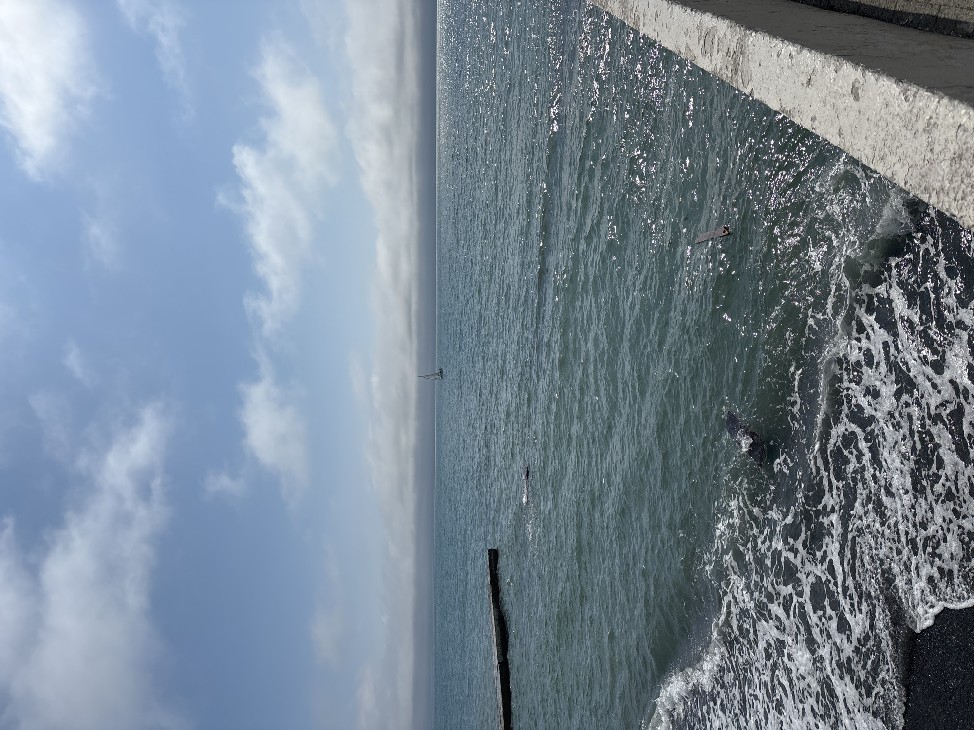 The image shows a seaside view, likely taken from an elevated position such as a pier or promenade, in Sochi, Russia. The scene includes a cloudy sky, a calm sea with a single sailboat visible in the distance, and an area where the sea meets a dark sand beach. Waves are crashing onto the beach, creating white foam. A weathered wooden post is partially submerged in the water, near the shoreline. In the top right corner of the image, there is a section of a concrete barrier or wall.