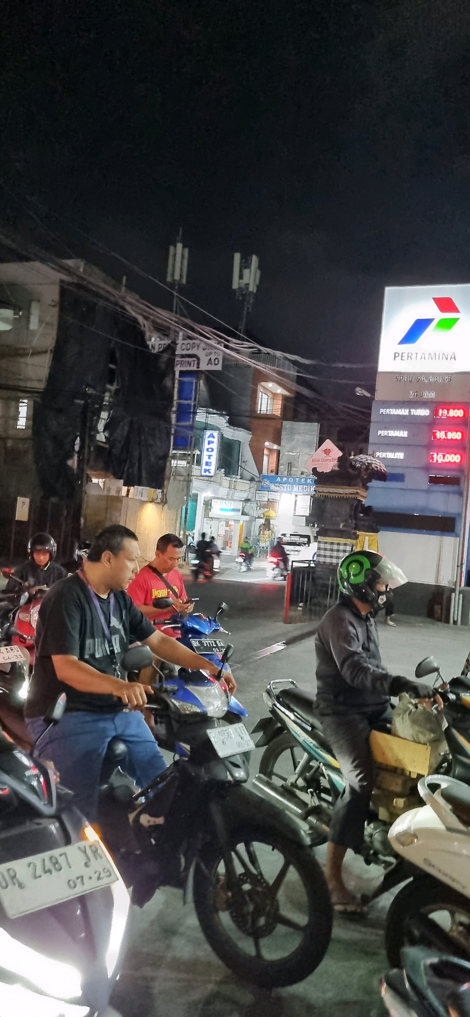 The image depicts a nighttime scene at a Pertamina gas station in Denpasar, Indonesia. The foreground features several individuals on motorcycles. In the center, a man wearing a black t-shirt and blue jeans is seated on a blue motorcycle, with his hand resting on the handlebar. To his right, another man in a red t-shirt is looking at his smartphone. Further to the right, a person wearing a helmet and dark clothing is on a motorcycle. In the background, the Pertamina signage is prominently displayed, showing fuel prices. Several other motorcycles and people are visible, indicating a busy gas station. The surrounding environment includes buildings with illuminated windows, power lines, and street lights, characteristic of an urban setting. License plates are visible on some of the motorcycles.