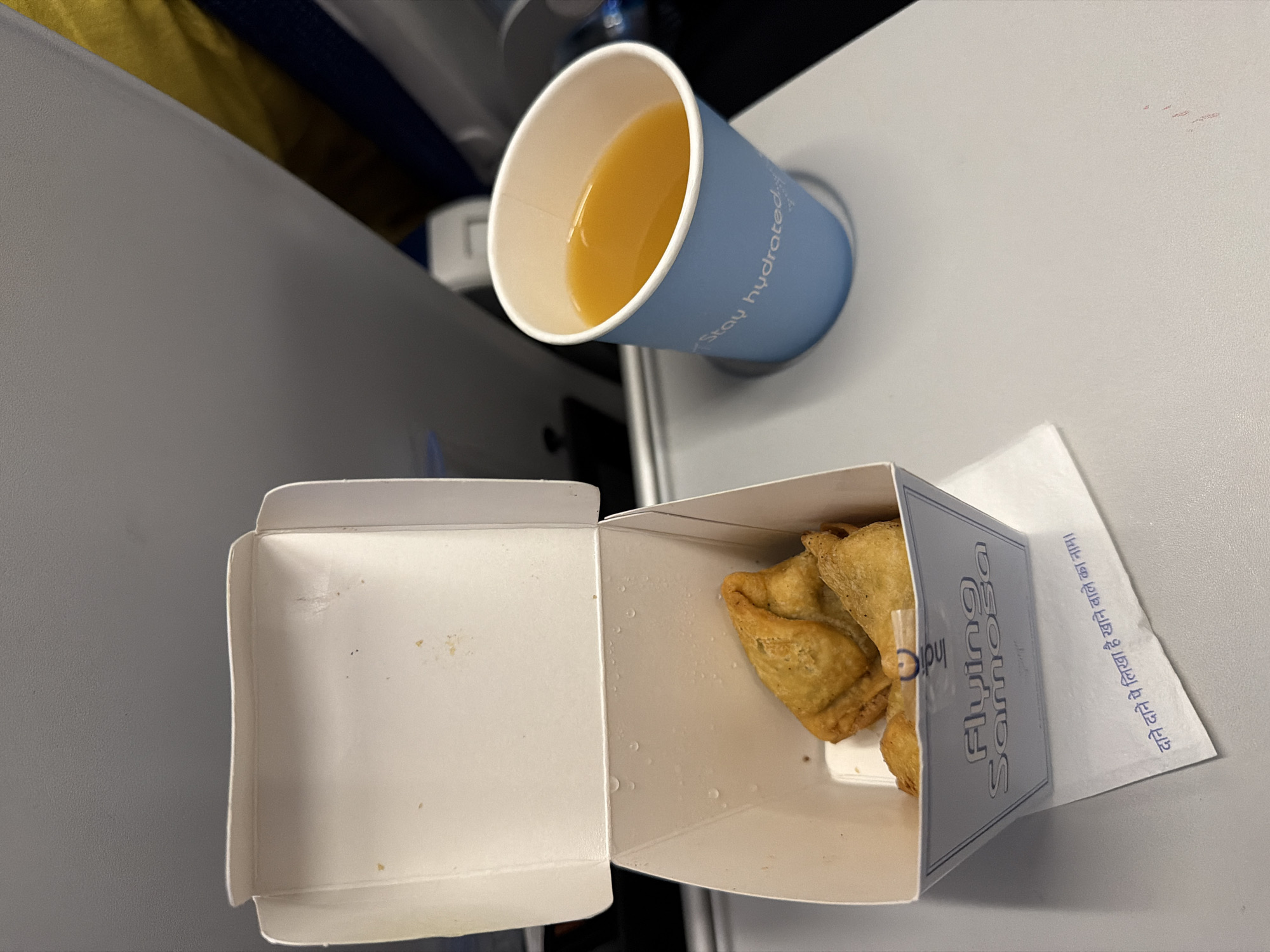 The image shows a tray table on an airplane with a box of samosas and a blue cup of orange juice. The box is white with "Flying Samosa" printed on it. There are three samosas in the box. The cup has "Stay hydrated" printed on it in white text. The tray table is gray and is set against a background of airplane seats. There is some text in Hindi on a napkin under the samosa box. The image was taken in Nadergul, India.