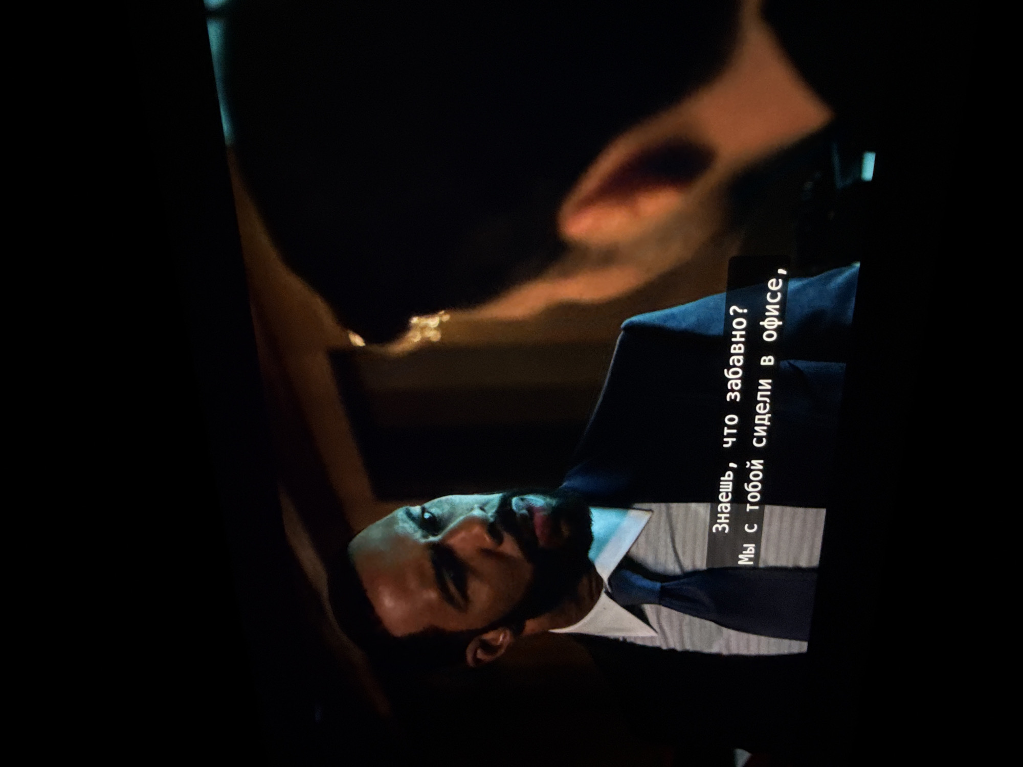 A medium shot presents a man with a beard and dark hair, facing left. He wears a white collared shirt, a dark tie, and a dark suit jacket. The image seems to be captured from a video screen, as there are subtitles in Russian on the right side that translate to "You know what's funny? We sat with you in the office". An out-of-focus figure looms above him. The image is dark, and the location of Troitsk, Russia is not discernable from the picture.