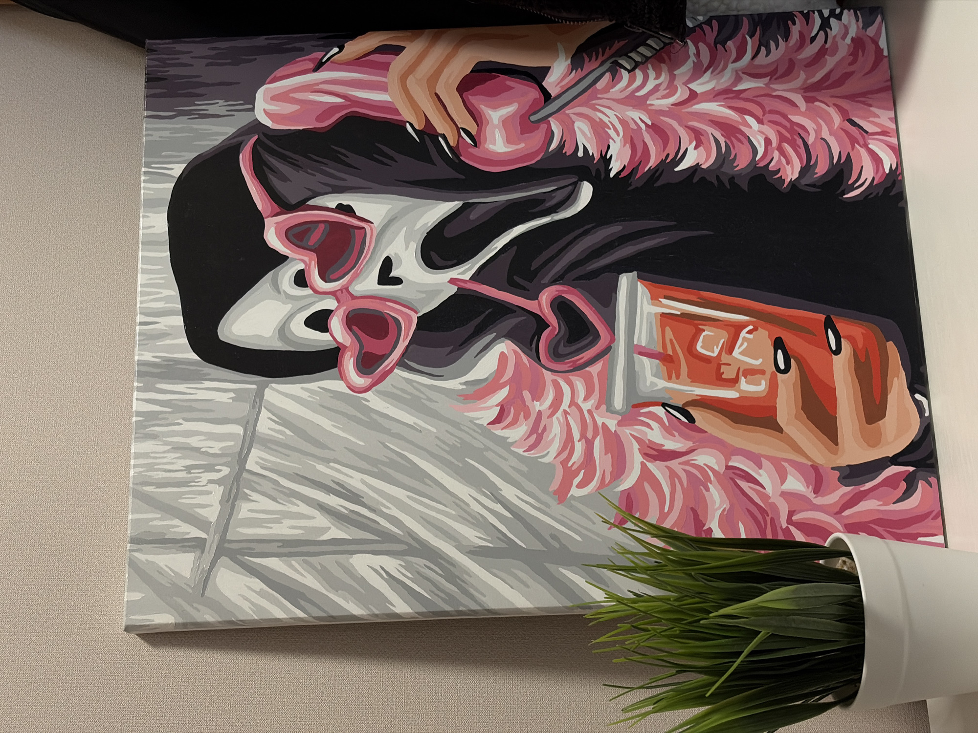 The image shows a painting hanging on a wall. The painting depicts a stylized female figure in a pool. She wears heart-shaped sunglasses and has a black bob haircut. A hand with black nail polish holds a lollipop. The figure is adorned with a pink feathered boa. There is an unidentified drink, likely a soda, behind the female. The background is stylized to resemble pool tiles. In the bottom right of the image is a white pot with green plastic plants. The scene does not show the city of Krasnodar, Russia.