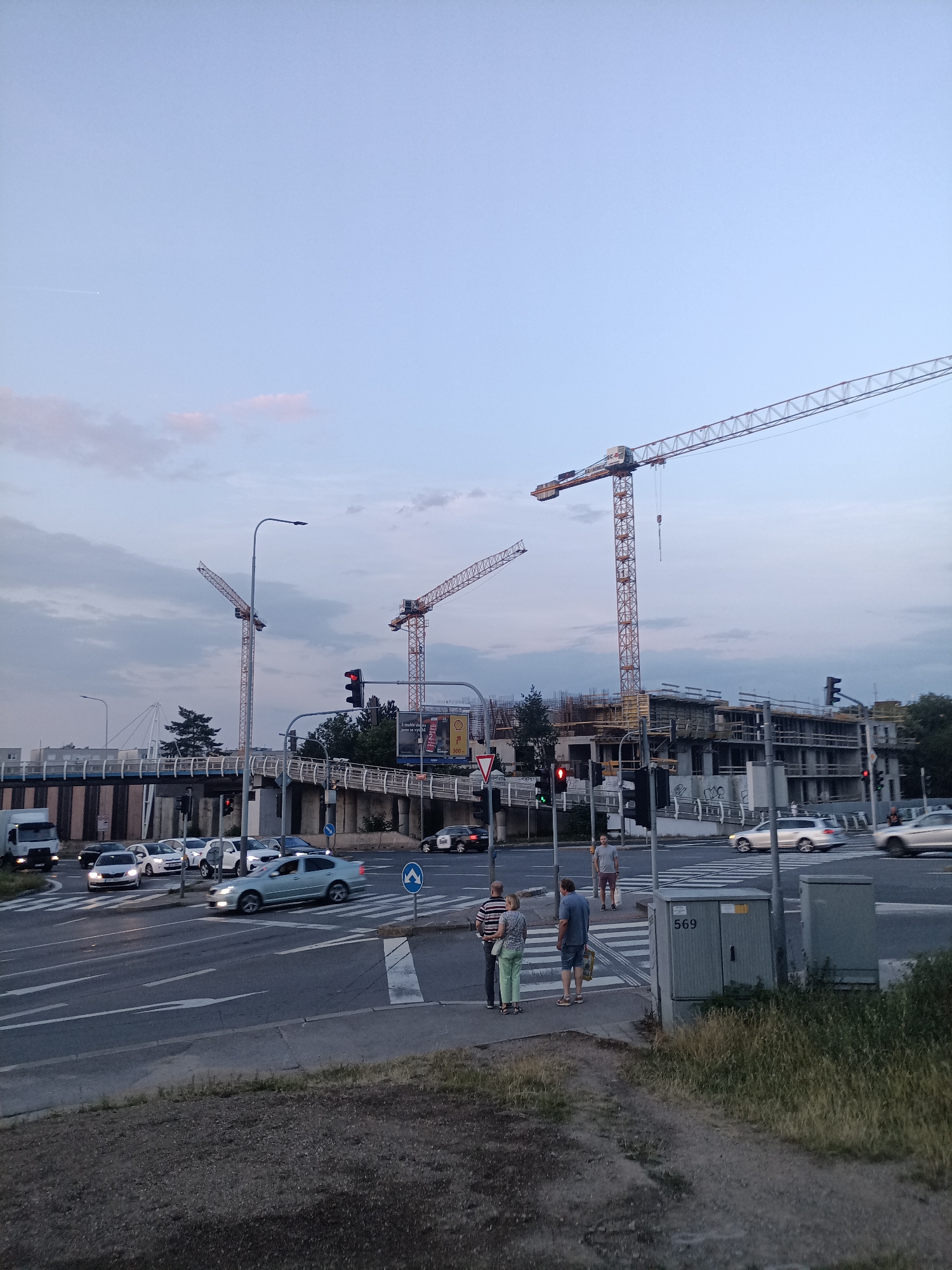 The image captures a street scene in Prague, Czechia, at dusk. Three prominent construction cranes dominate the skyline, indicating ongoing development. A multi-level roadway or overpass is visible in the background, with cars traversing it. In the foreground, a crosswalk is occupied by several pedestrians. Cars are also present on the road, some in motion, suggesting a busy intersection. The sky is a muted blue with hints of pink from the setting sun, and a faint contrail can be seen. The overall atmosphere is one of urban activity under a fading light.