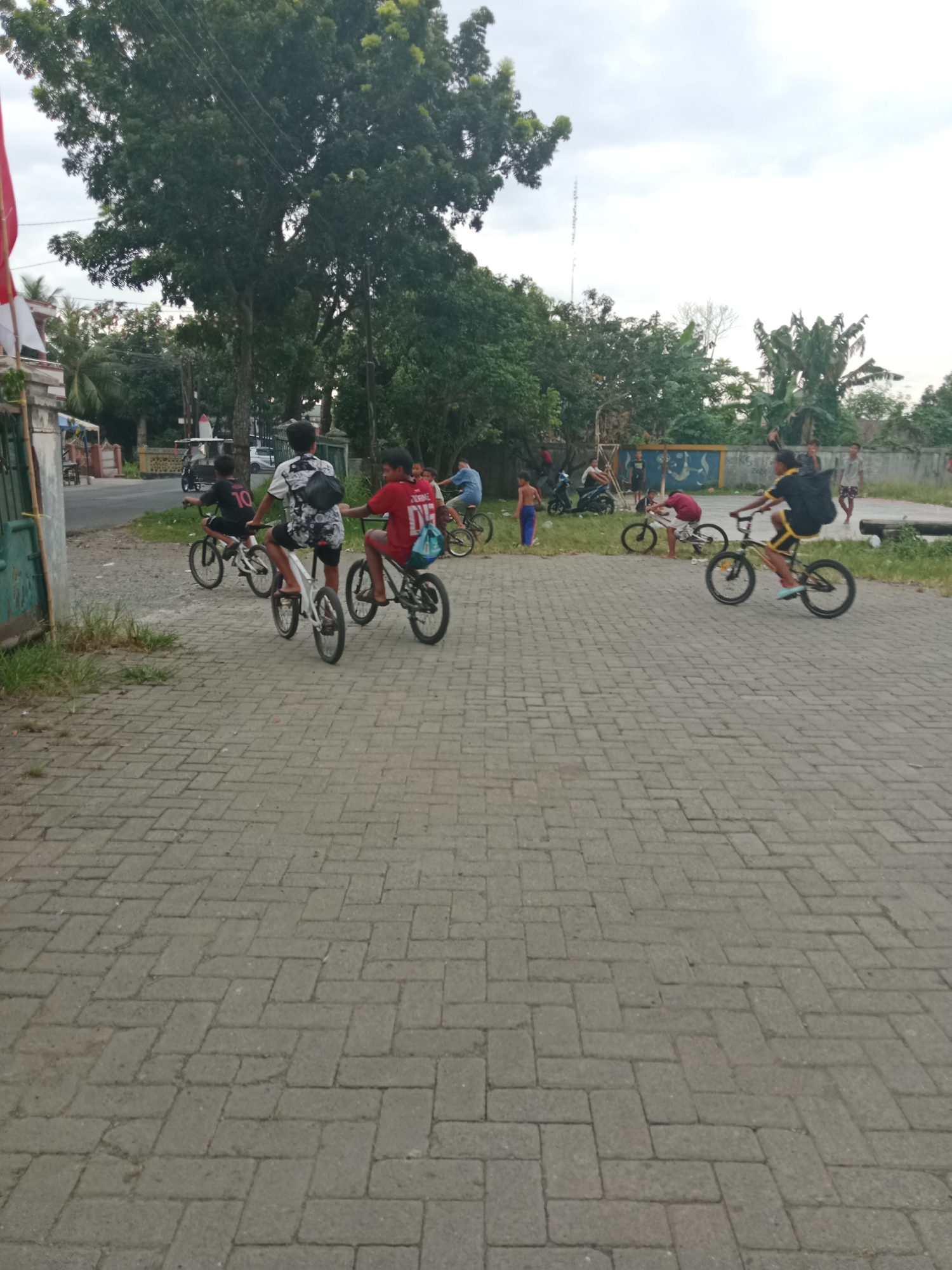 A group of young males are riding bicycles on a paved walkway in what appears to be a residential area of the City of Binjai, Indonesia. In the foreground, a young male wearing a black t-shirt with yellow trim and a backpack rides a dark-colored BMX bicycle. Behind him, three other boys on bicycles are also visible, including one in a red shirt and another in a black shirt with the number 10 on the back. The walkway is made of interlocking gray brick pavers. To the left of the walkway is a dirt shoulder with green grass and a few small trees. Beyond that, there is a street with a few parked motorcycles and a building with a red and white flag. In the background, there are more trees and a wall with a mural. The sky is overcast.