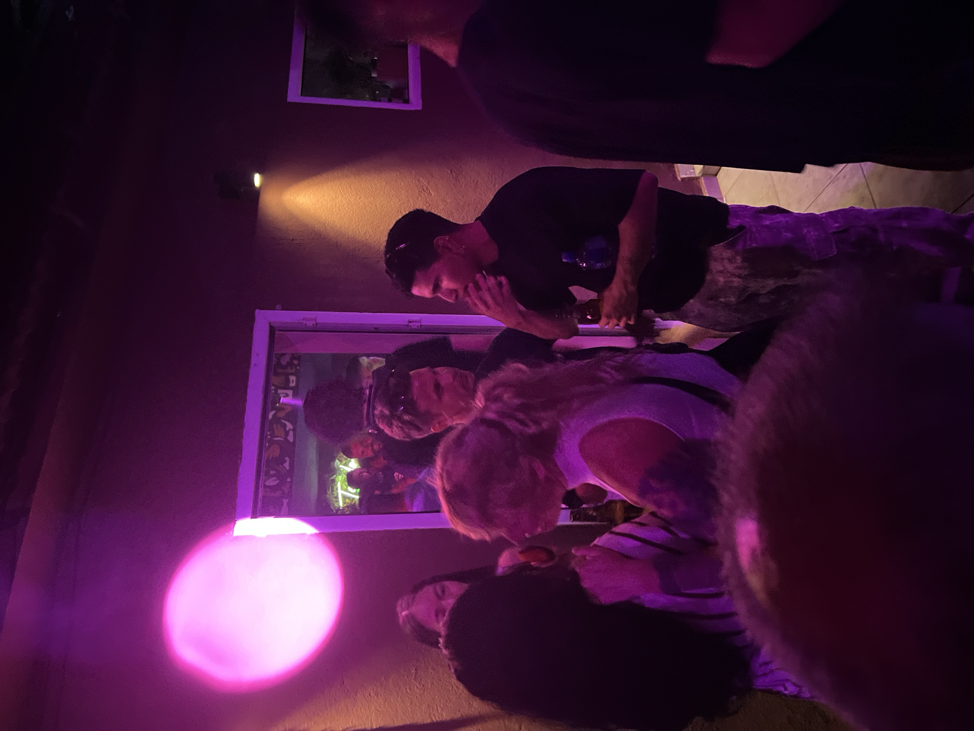 The image depicts a dimly lit interior, likely a bar or club, with a prominent pink light source illuminating the left side of the frame. Several individuals are visible, engaged in social interaction. A person in the foreground, on the left, has dark hair and is partially obscured. To their right, a person with blonde hair is looking towards the left, wearing a light-colored top. Further back, a man in a black t-shirt is holding a bottle and appears to be speaking or listening, with his hand near his mouth. His attire includes camouflage-patterned pants. Another man, with short dark hair and sunglasses on his head, is visible behind him. The background reveals a doorway or entrance with reflections and a glimpse of another room, possibly with plants. The overall ambiance suggests a social gathering in an urban setting, potentially in Herceg Novi, Montenegro, given the context. The lighting is atmospheric, with a mix of pink and orange hues.