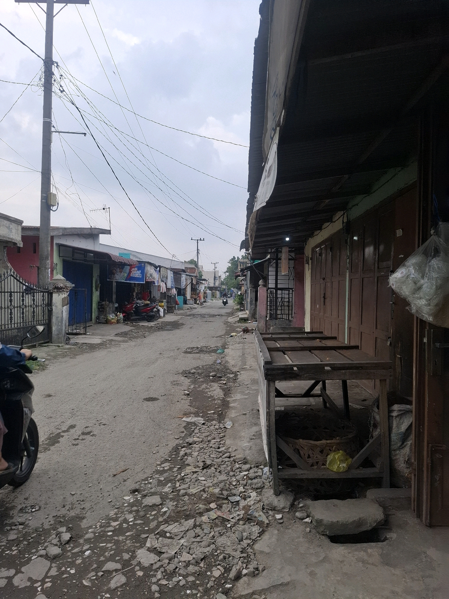 The image depicts a narrow, unpaved street in the city of Kenangan, Indonesia, lined with small shops and residential buildings. The street is littered with debris, including rocks, trash, and puddles of water. Overhead, a tangle of electrical wires crisscrosses the sky, supported by a prominent utility pole on the left.
On the left side of the street, a black metal gate stands in front of a building with red and gray facades. A scooter, partially visible, is parked near the gate. Further down the street, more shops are visible, some with open fronts displaying goods and others with closed, wooden doors. Motorcycles are parked in front of some of these establishments. A sign with Indonesian text, "JADI JUARA! Mizone," is prominently displayed above one of the shops.
On the right side of the street, a wooden stall with empty shelves sits in front of a row of shops with closed, brown wooden doors. A large, clear plastic bag filled with white objects hangs from one of the shop fronts. A woven basket containing a yellow plastic bag is visible beneath the wooden stall. The ground in front of the shops is a mix of concrete and dirt, with a large, flat stone covering a hole in the ground.
The sky is overcast with a pale, diffused light. The overall impression is one of a modest, working-class urban environment with signs of daily life and commerce.