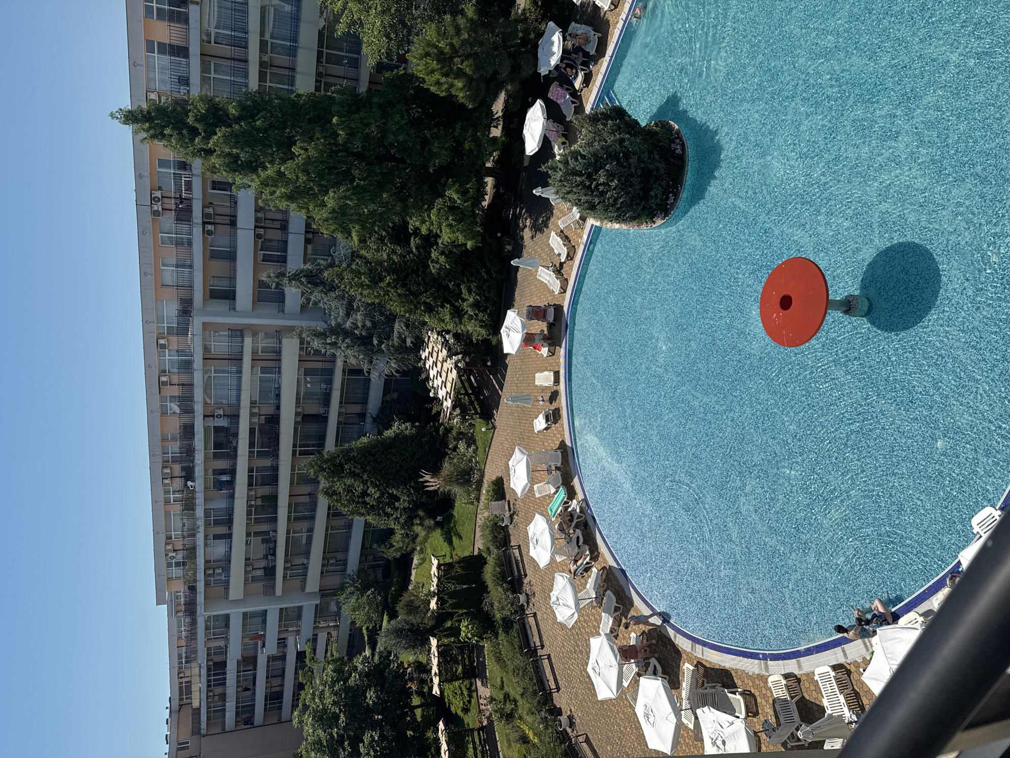 An aerial view captures a resort swimming pool area in Nesebar, Bulgaria. The large, irregularly shaped pool is filled with clear blue water, featuring a red mushroom-shaped table in the center. Surrounding the pool is a paved area with numerous white lounge chairs and umbrellas, some occupied by people. To the left, a multi-story hotel building with balconies and air conditioning units is visible, with trees and greenery in front of it. The sky is clear and blue.