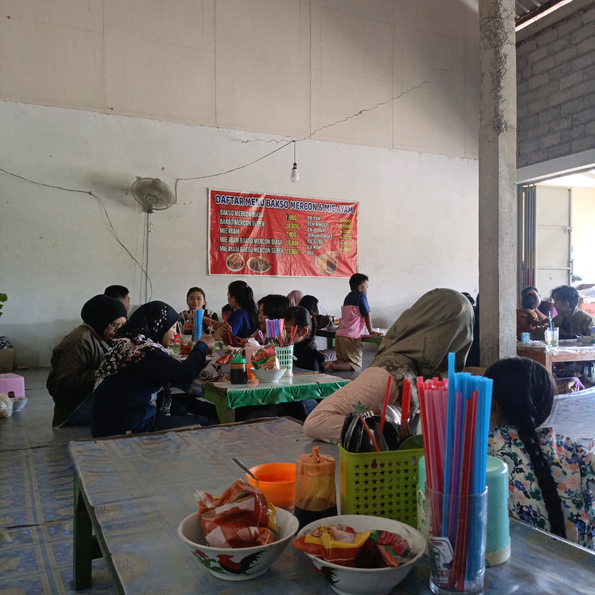 The image depicts an indoor eating establishment, likely a warung or small restaurant, in Salatiga, Indonesia. Several people are seated at tables, engaged in eating and socializing. The tables are covered with blue plastic cloths, and food items such as bowls of what appears to be noodles or soup, sauces, and drinks are visible. A red banner displaying a menu in Indonesian is hung on the wall, listing "BAKSO MERCON & MIE AYAM" with prices. The environment appears somewhat rustic, with exposed concrete pillars and a tiled floor. A ceiling fan is mounted on the wall, and a light bulb hangs overhead. The lighting suggests it is daytime. People of various ages, including children and adults, are present, some of whom are wearing headscarves, indicating a predominantly Muslim population. The overall scene conveys a casual dining experience in a local Indonesian setting.