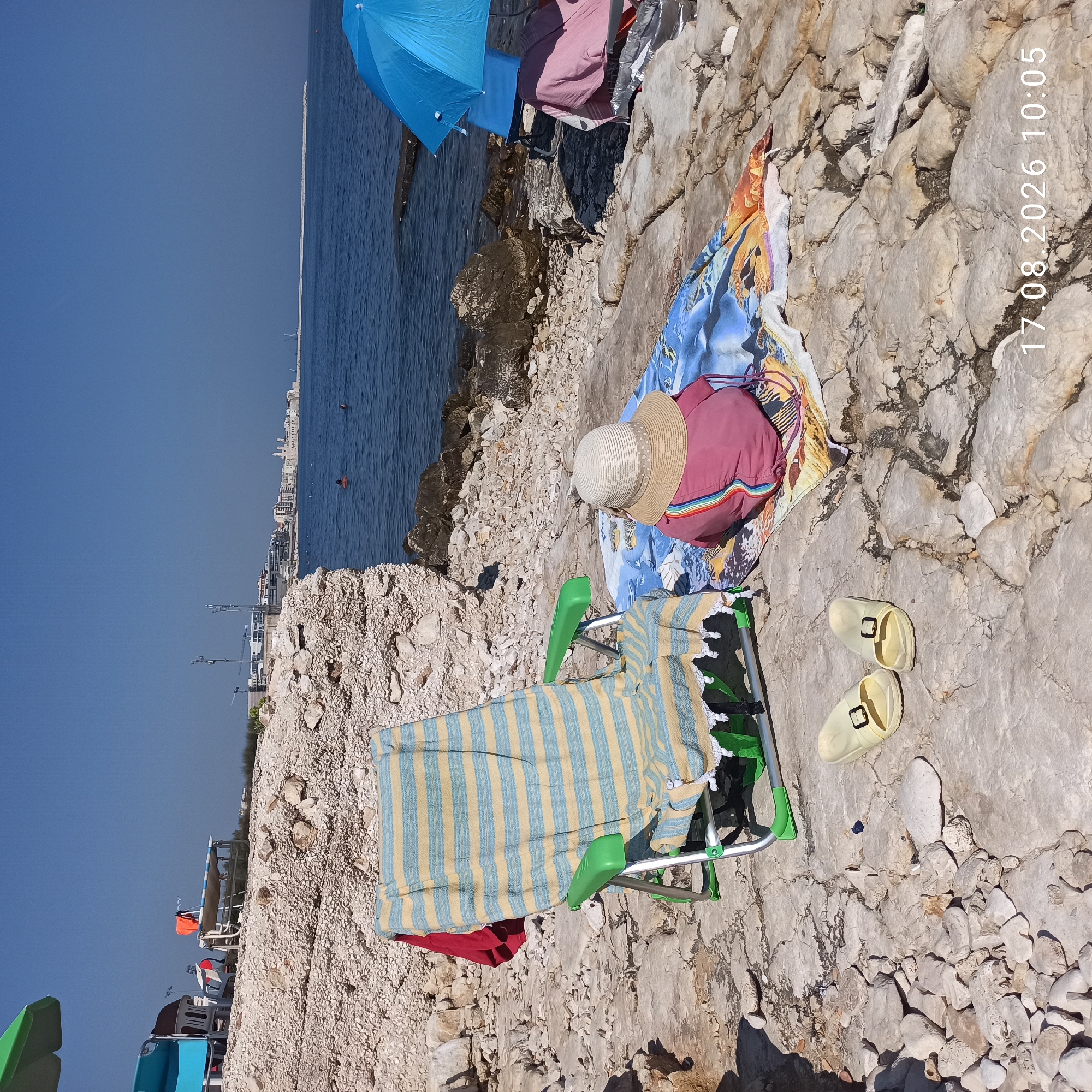 The image depicts a rocky beach in Molfetta, Italy, under a clear blue sky. In the foreground, a striped beach chair with a yellow and blue towel is set up on the stony ground. Beside the chair, a child wearing a pink shirt and a straw hat sits on a patterned beach towel, facing the sea. A pair of yellow flip-flops rests on the rocks near the chair. Further back, the blue Mediterranean Sea stretches out, with a distant cityscape visible on the horizon. A large blue umbrella and a red towel are placed on another chair to the right of the frame. The environment suggests a leisurely day at the beach, with the timestamp "17.08.2026 10:05" indicating the time the photo was taken.