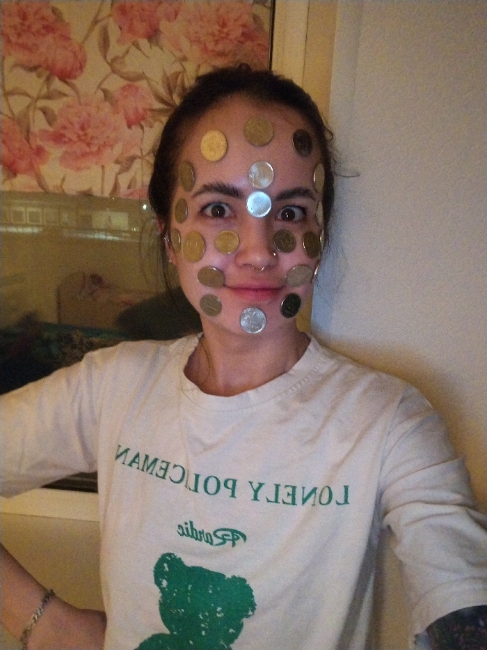 A female individual is depicted in a medium shot, indoors. Her face is extensively covered with approximately 25-30 circular metallic coins of various diameters and hues, including silver, gold-toned, and bronze-toned, affixed to her forehead, cheeks, nose bridge, upper lip area, and chin. Her dark hair is pulled back, and her eyes are wide open, gazing directly forward. A small, silver-toned ring pierces her left nostril. She is wearing a light-colored, long-sleeved t-shirt. The shirt features a green, mirrored graphic displaying the text "NAMECILOP YLENOL" (which reads "LONELY POLICEMAN" when un-mirrored) above a green silhouette of a teddy bear. A metallic link bracelet is visible on her left wrist. The background on the left side of the frame features a window or reflective panel, partially revealing a light-colored surface with a pink floral (peony-like) pattern, possibly a curtain or wallpaper, along with faint reflections of what appear to be interior or exterior lights. The background on the right side is a plain, light-colored wall. The scene is located in the city of Noviy, Russia.