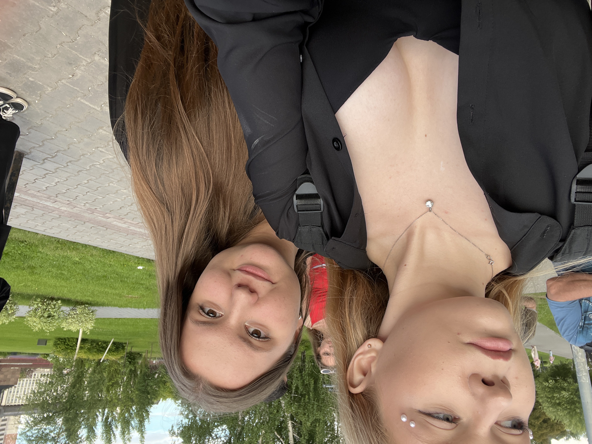 In the foreground of the image, two young women are positioned close together. The woman on the left is partially obscured, facing away from the camera and looking to her left. She has blonde hair and is wearing a black top and a black unbuttoned shirt, with a silver pendant necklace. Several small pearl-like decorations are affixed to her forehead and eyebrow. The woman on the right is facing the camera, with long brown hair. She is wearing a black garment. Behind them, a park-like setting is visible with trees and green grass. In the background, other people are present, including a person wearing a red top. The ground appears to be paved with interlocking grey bricks. The overall lighting suggests it is daytime. This scene is likely in the city of Perm, Russia, given the context of the location being provided.