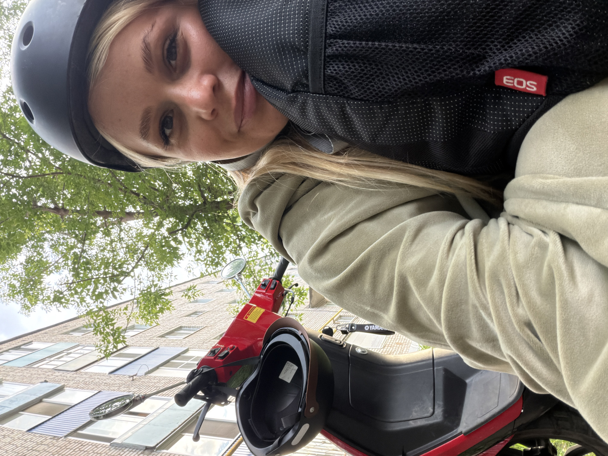 A young blonde woman, wearing a black helmet and a light green hoodie, is captured in a close-up selfie next to a red scooter. She is resting her head on a black backpack, which has a small red tag with white lettering that reads "EOS". The scooter has a black helmet hanging from its handlebars. In the background, a brick apartment building with white-framed windows is visible, along with green trees and a cloudy sky. The scene appears to be on a street in Sarapul, Russia.