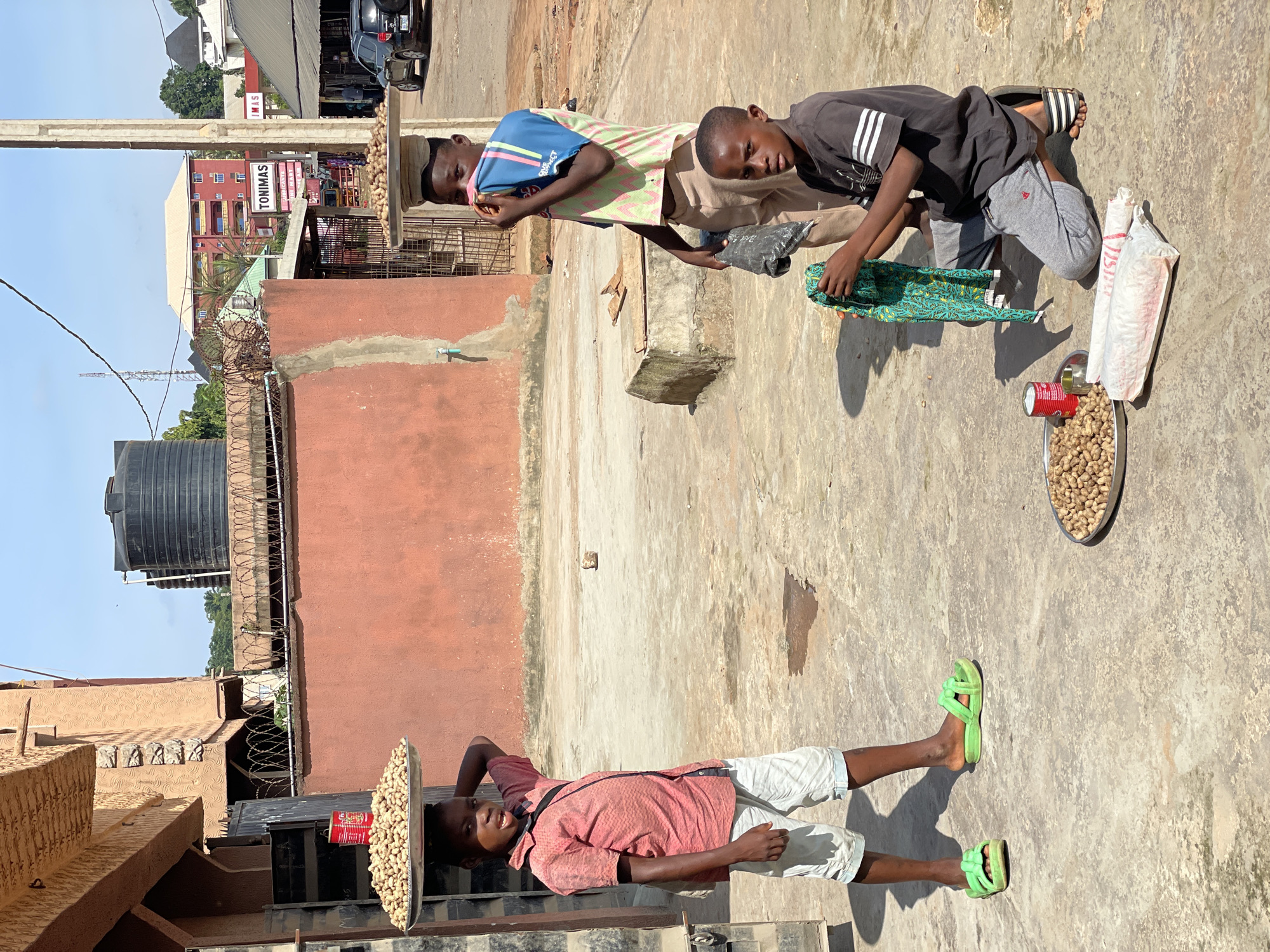 In Nkpor, Nigeria, three young boys are seen outdoors under a clear, bright sky, suggesting it is a sunny day. The scene appears to be a marketplace or a street with buildings and makeshift stalls.
The main subjects are the three boys, likely teenagers or pre-teens. One boy, wearing a pink shirt and white shorts, stands with a tray of nuts balanced on his head. He is looking to the side and has his right arm casually behind his head. Beside him, another boy in a blue and green striped shirt and light brown shorts is holding what looks like a folded piece of fabric. He is looking down and appears to be in conversation or interaction with the boy next to him. This third boy is wearing a dark t-shirt with white stripes on the sleeves and grey shorts. He is crouched down, also holding a colorful piece of fabric, possibly to display or sell. In front of him, on the ground, is a tray filled with nuts, a red can, and two white plastic bags.
The ground is paved with concrete, and there are elements of the urban environment visible, including a black water tank, barbed wire fencing, and buildings with red and beige facades. A sign with the word "TONIMAS" is visible on one of the buildings. The overall impression is a moment of everyday life, likely related to commerce, given the display of goods. The boys' expressions are neutral, suggesting a typical day rather than a special event. The sunlight casts distinct shadows, indicating it is likely midday.