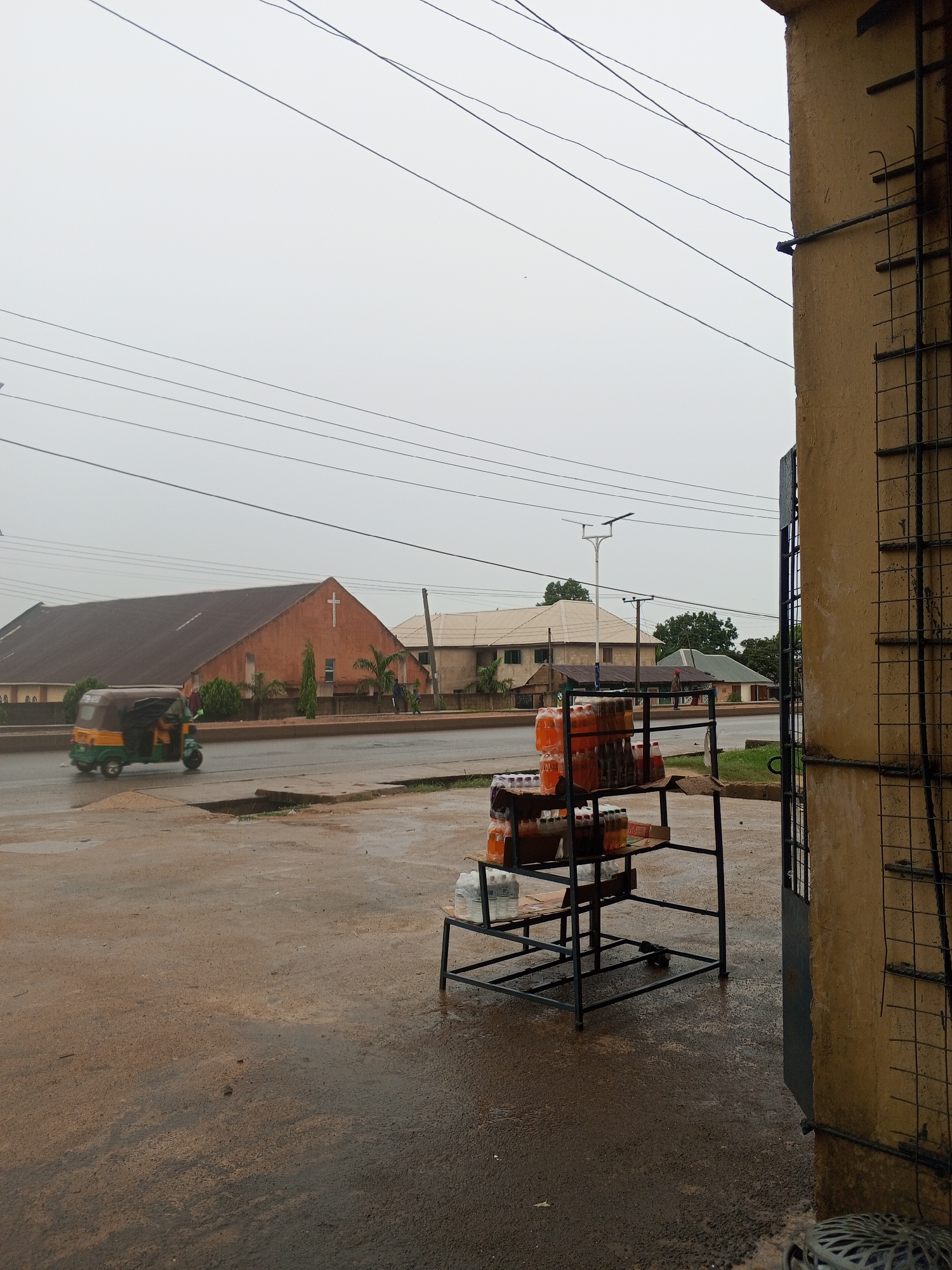 The image shows a wet street scene in Unknown, Unknown. The sky is overcast. On the left, a green and yellow auto rickshaw is driving on the road in front of a rust-colored building with a cross on its facade. Further down the road is a tan building. In the foreground, to the right, there's a metal rack with bottles of orange soda and clear water bottles stacked on it. Part of a building with a metal grate on its side is visible on the right edge of the frame. The street and sidewalk are wet, indicating recent rain. Power lines run across the sky.