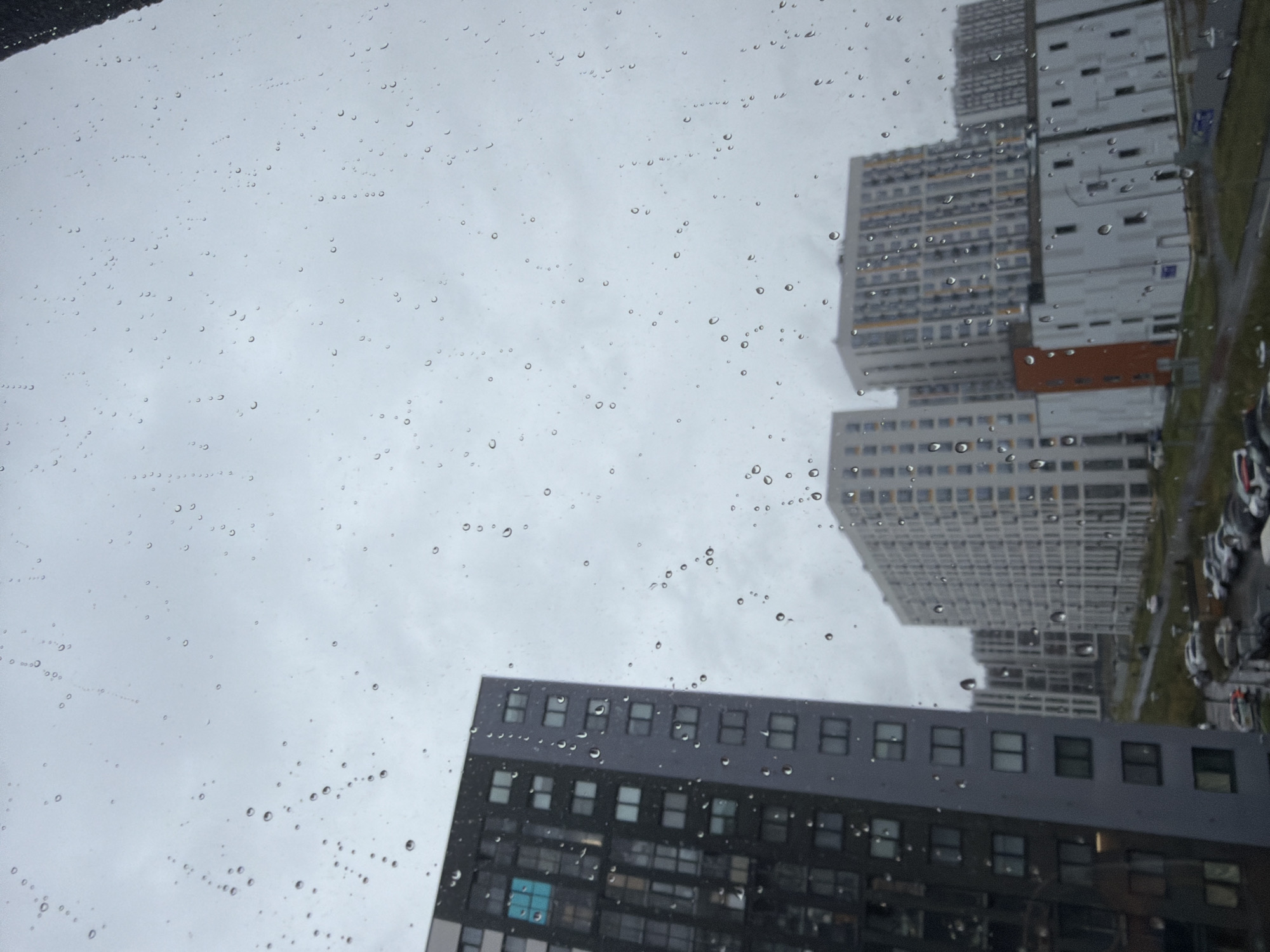 The image depicts a view of a residential area in Pargolovo, Russia, through a window covered in raindrops. In the foreground, a tall, dark grey apartment building with numerous windows is visible on the left. To its right and in the background, several other apartment buildings of varying heights and architectural styles are seen. These buildings are predominantly white and grey, with some featuring yellow accents. The sky is overcast and grey, suggesting rainy weather. Below the buildings, a road with parked cars can be observed. The presence of raindrops on the window creates a slightly blurred and textured effect, obscuring some of the finer details of the scene. There are no people visible in the image.