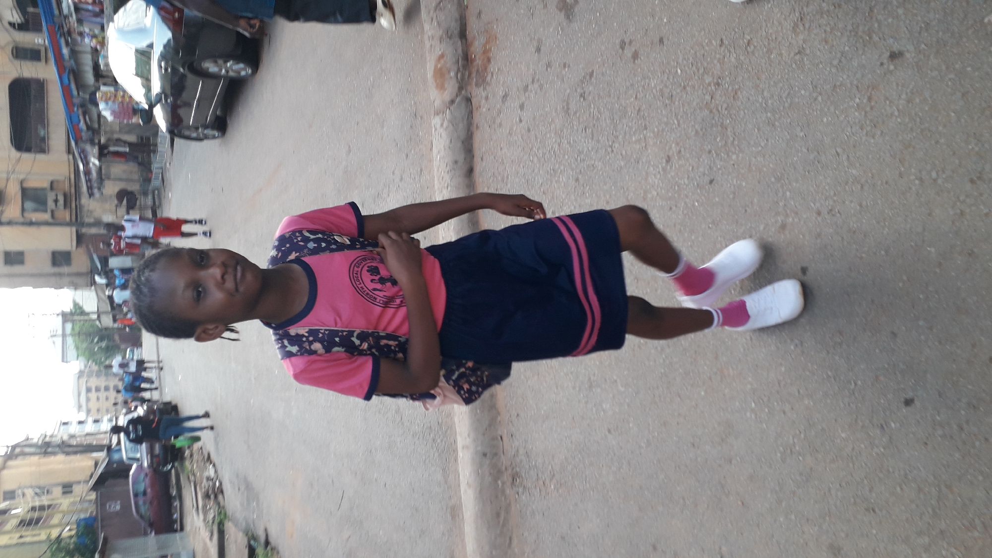 The image depicts a young girl in a pink and blue school uniform with white shoes and pink socks, standing on an asphalt street. She has a backpack and is smiling. The background includes buildings, people walking, and cars. The scene is an urban street in Onitsha, Nigeria.