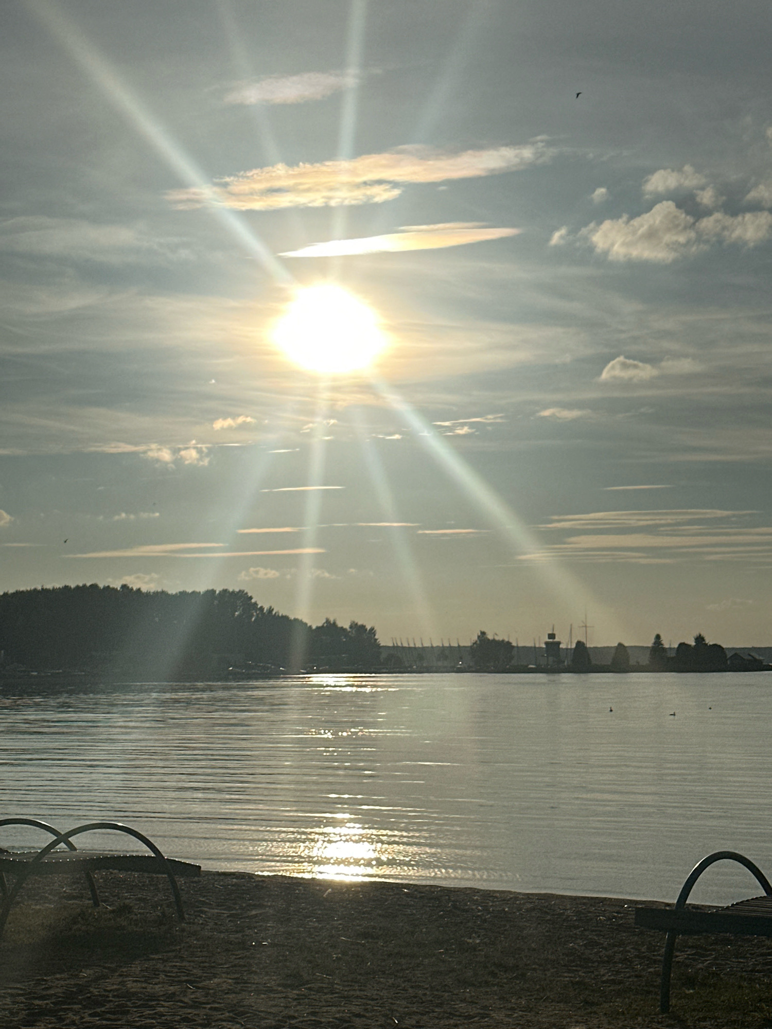 This image depicts a serene lakefront scene, likely during sunset or sunrise, with the sun's rays prominently visible and creating a lens flare effect across the sky. The foreground shows a sandy beach with two empty lounge chairs. The middle ground features a calm body of water reflecting the sunlight, with a silhouetted treeline on the far bank, interrupted by structures resembling a marina with several sailboats and a lighthouse or tower. The sky is partially cloudy, with thin, wispy clouds and some larger, puffy clouds. A small bird is visible in flight against the sky. The scene evokes a peaceful, tranquil atmosphere. The location is identified as Zhdanovichsky Sel'skiy Sovet, Belarus, suggesting this is a view of the Minsk Sea (Zaslavskoye Reservoir) or a similar body of water in that region.