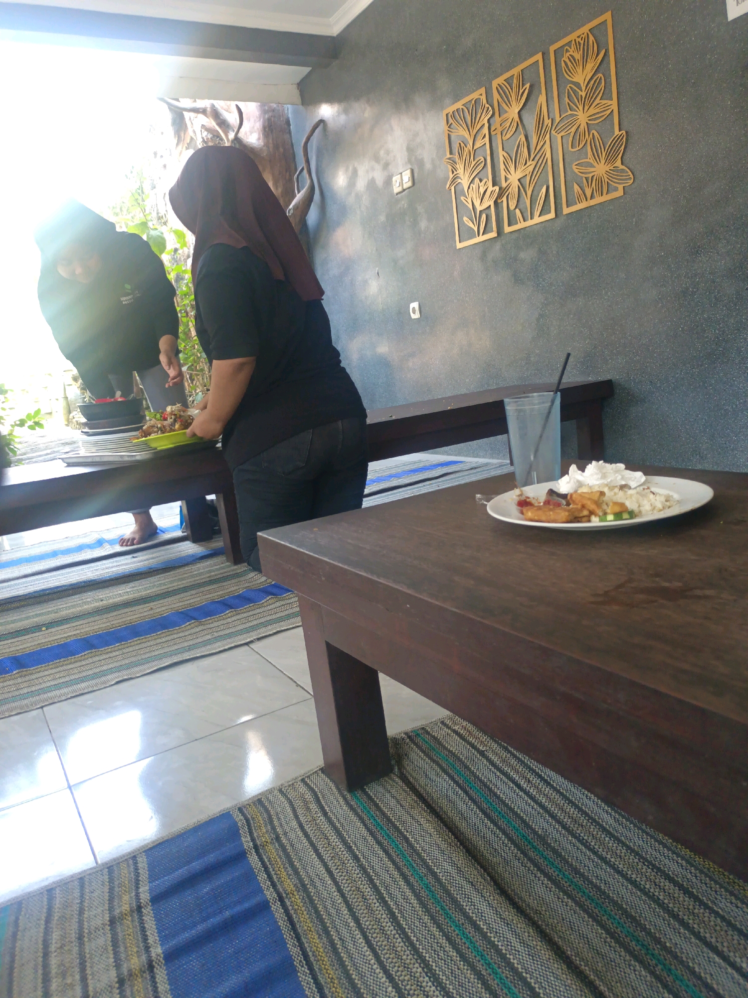 In the city of Kerangkulon, Indonesia, a dimly lit outdoor seating area features two individuals by a low wooden table. One person, wearing a dark headscarf and black clothing, is placing a plate of food on the table. Beside them, another person in a dark hoodie is looking down. In the foreground, a second dark wooden table holds a plate with food, a glass of water, and a straw. The ground is covered with striped mats. The wall behind the individuals is decorated with three golden floral wall hangings.