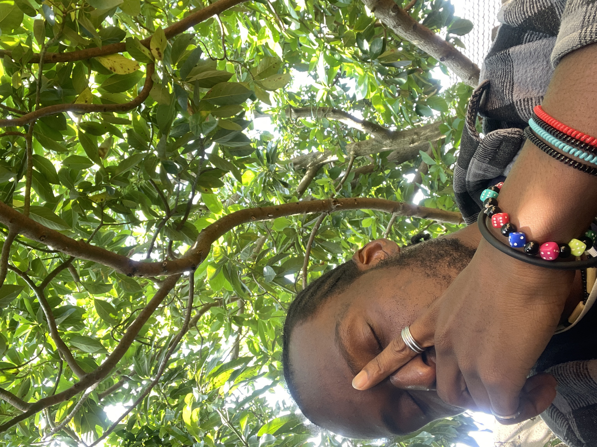 The image is a close-up, low-angle shot of a Black individual with their eyes closed, resting their forehead and nose with their right hand. The individual is wearing several rings on their right hand, including two silver bands on the index finger. They are also wearing bracelets on their wrist, one with colorful dice-shaped beads and another with black, teal, and red beads. The individual is wearing a plaid shirt. The background consists of a tree with lush green leaves and branches, with sunlight filtering through the foliage. The scene appears to be outdoors, possibly in a garden or park. The general environment suggests a warm, possibly tropical or subtropical climate. There are no immediate indicators to pinpoint the exact location within Jos, Nigeria, but the natural setting is consistent with areas in and around the city.