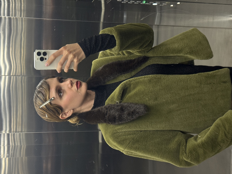 A young woman takes a selfie in an elevator. She wears an olive green faux fur coat with a dark brown fur collar, and a black top with sparkly sleeves. Her blonde hair is styled in waves with a silver clip. She holds a white smartphone in her left hand, focusing on the camera. The elevator walls are metallic and shiny.