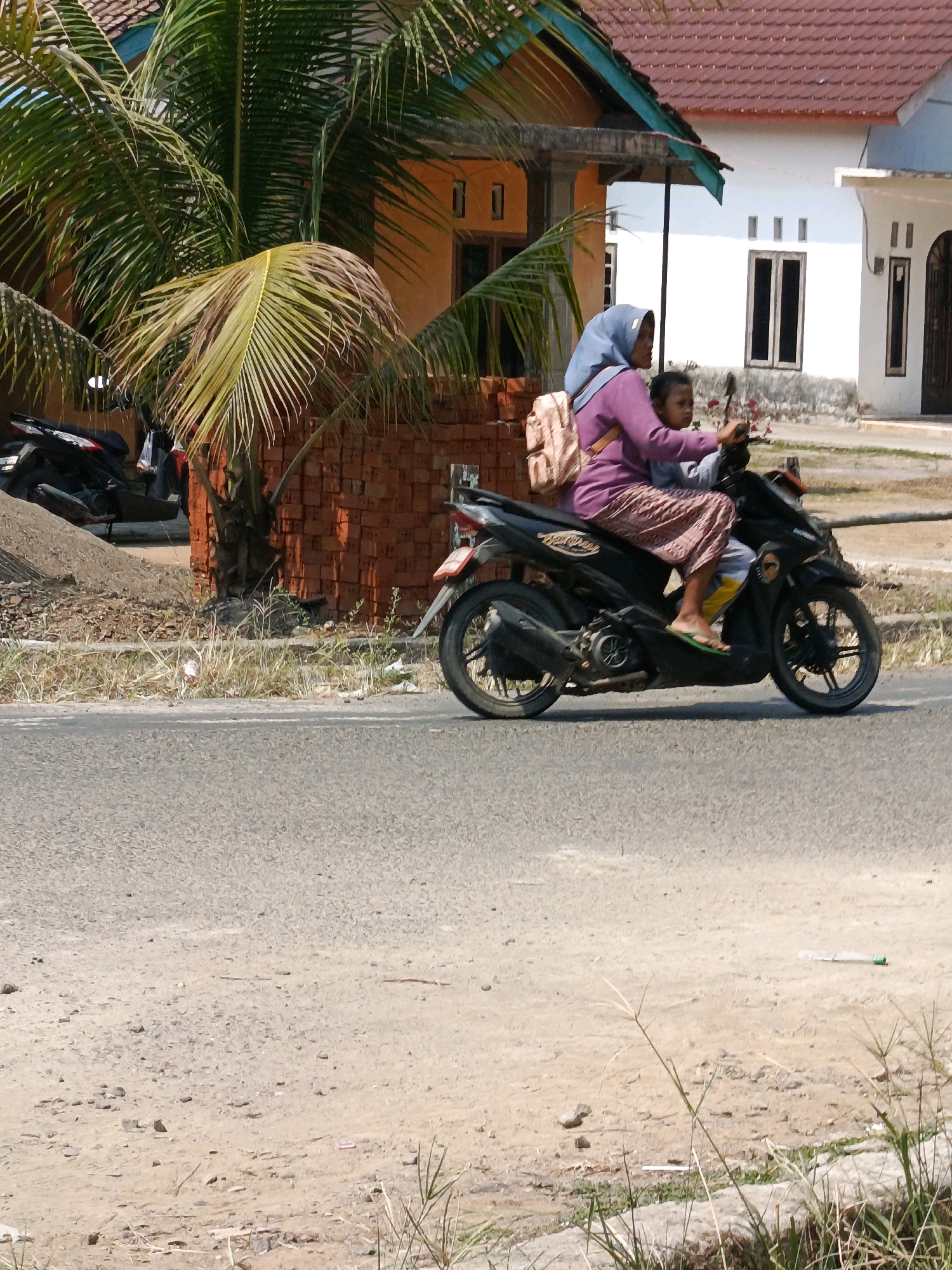 In Wonosari, Indonesia, a woman wearing a blue hijab and a purple top is riding a black motorcycle. She is carrying a child in front of her, who is wearing a gray shirt and yellow shorts. The woman has a beige backpack slung over her shoulder. They are riding on a paved road, with a sandy area to the right containing dry grass and some debris. Behind them, there is a brick wall, a palm tree, and houses with tiled roofs. To the left of the woman, there are parked motorcycles. The scene appears to be a residential area on a sunny day.