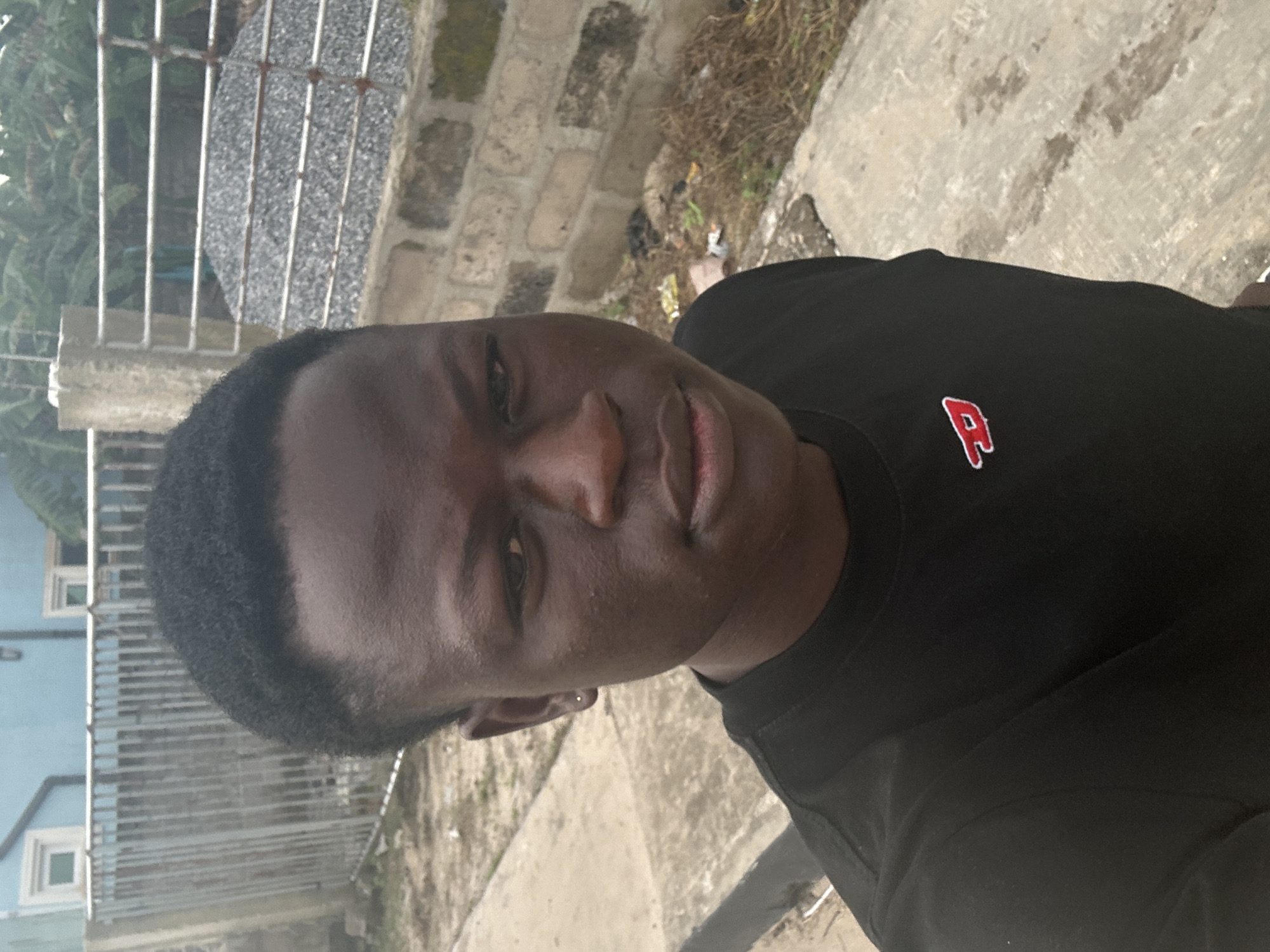 A young Black male wearing a black t-shirt with a red logo is looking directly at the camera. He has short, dark hair and is wearing a small gold stud earring in his left ear. The background suggests an outdoor setting in Badagry, Nigeria, with a light blue building, a metal fence, a brick wall, a pile of gravel, and some vegetation visible. The ground in the foreground appears to be concrete or pavement.