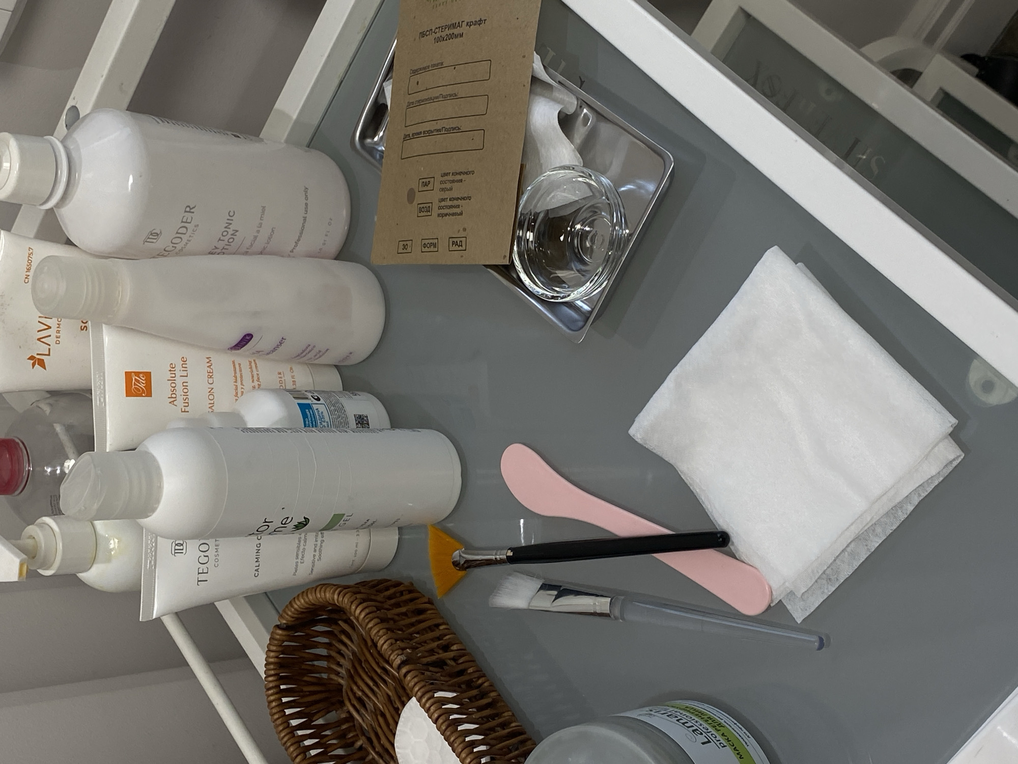 Here is a detailed description of the image:
**Scene:** The image shows a collection of skincare and cosmetic products neatly arranged on a gray surface, likely a tray or countertop.
**Objects:**
* **Skincare Products:** There are several bottles and tubes of skincare products, including toners, cleansers, creams, and gels. Prominent brands visible are "TEGODER," and "LAVI DERMO".
* **Cosmetic Tools:** A pink spatula, a cosmetic brush with a yellow tip, and another brush with clear plastic handle are present.
* **Paper Products:** There is a stack of white paper wipes. A brown paper card "ПСП-СТЕРИМАГ крафт" is also visible.
* **Glassware:** A clear glass bowl rests on a tray.
* **Basket:** A woven basket is present towards the bottom left, holding what appears to be cotton balls or other small cosmetic items.
* **Product containers**: a white bottle with gray label and a white lid.
**Environment:** The environment appears to be a clean, organized space, possibly a beauty salon, spa, or a makeup artist's workspace. The setting suggests a focus on beauty and skincare procedures.
**Details & Summary**
Overall, the image suggests a setup for facial treatments or cosmetic applications, with various skincare products and tools readily available.