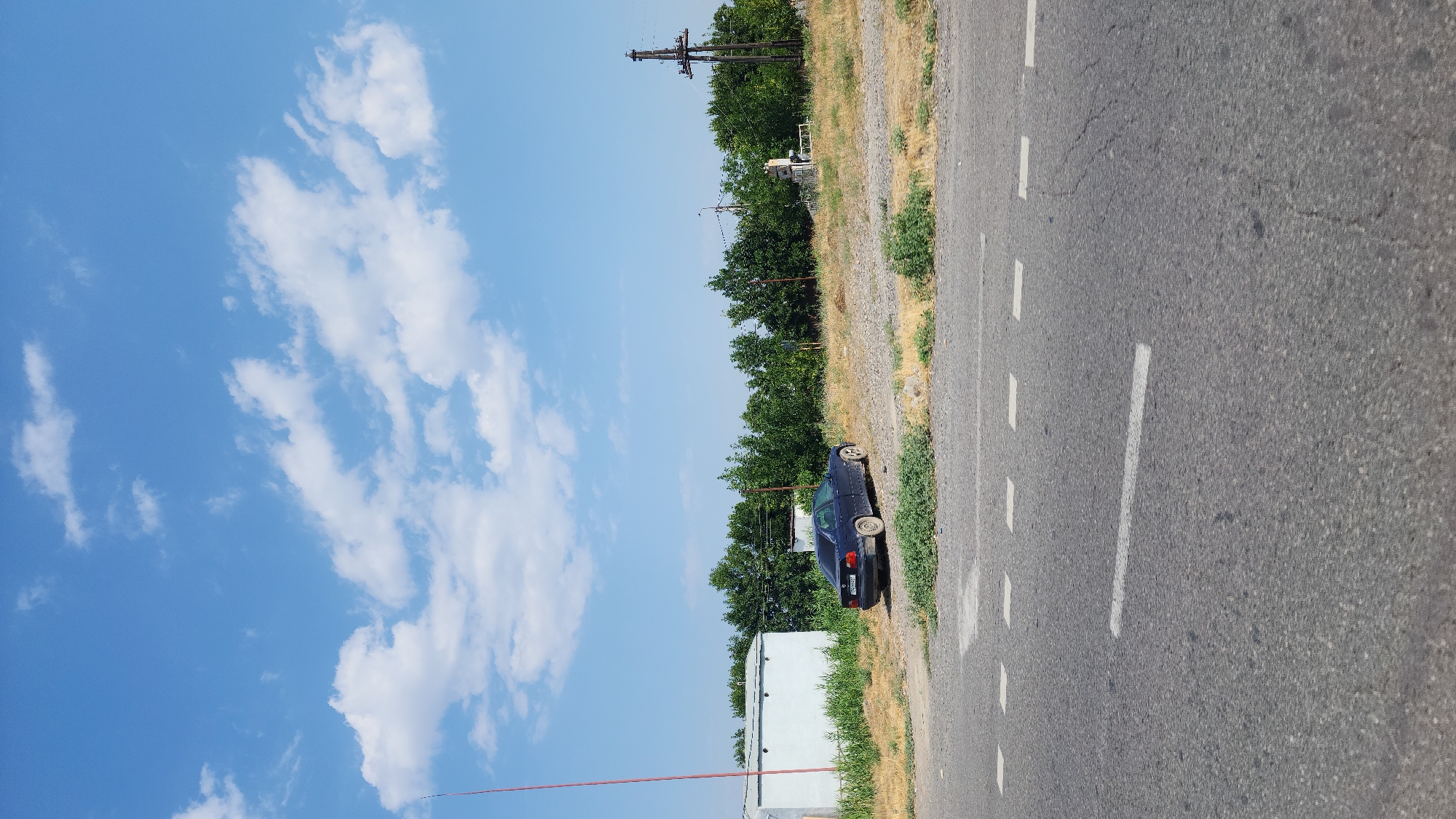 A dark sedan is parked on a dirt shoulder next to a road in Arshaluys, Armenia. The car is facing away from the camera and is slightly dusty. In the background, there is a white building to the left and a line of trees. A utility pole with electrical wires is visible in the distance on the right. The sky is blue with scattered white clouds. The foreground shows the asphalt road with white dashed lines indicating lanes.