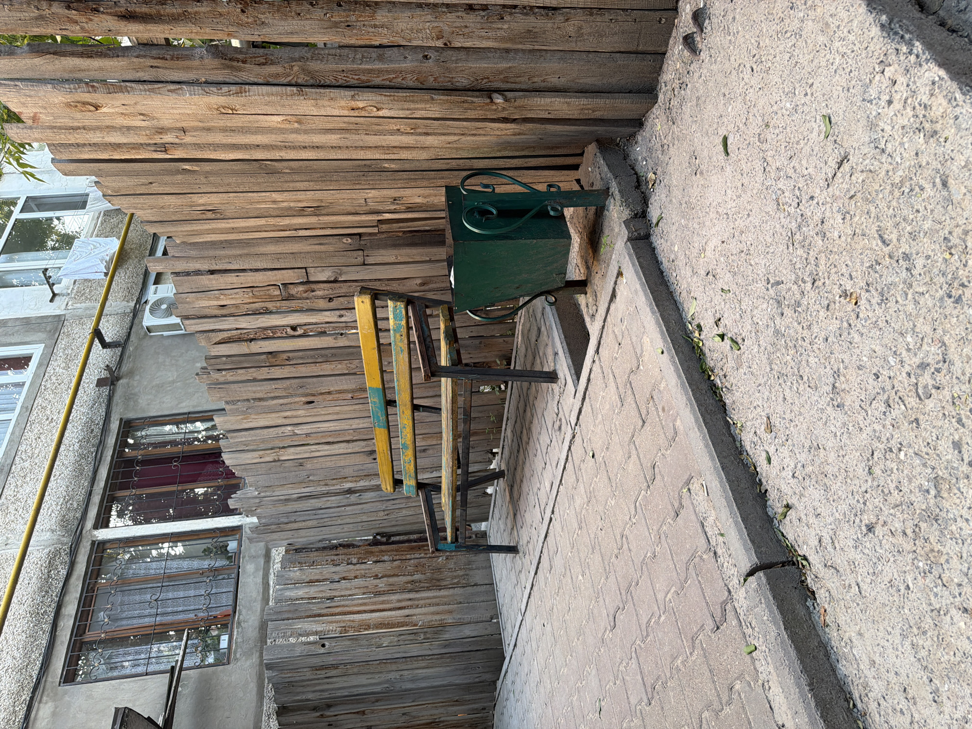 An exterior shot taken in Shymkent, Kazakhstan, shows a weathered wooden fence partitioning a courtyard area. To the left, a section of a building with barred windows is visible. In the foreground, a paved area transitions to concrete. A bench with a yellow and green painted seat and black metal legs stands on the paved section, adjacent to a green metal trash can with decorative metalwork. The scene is bathed in natural light, suggesting daytime.