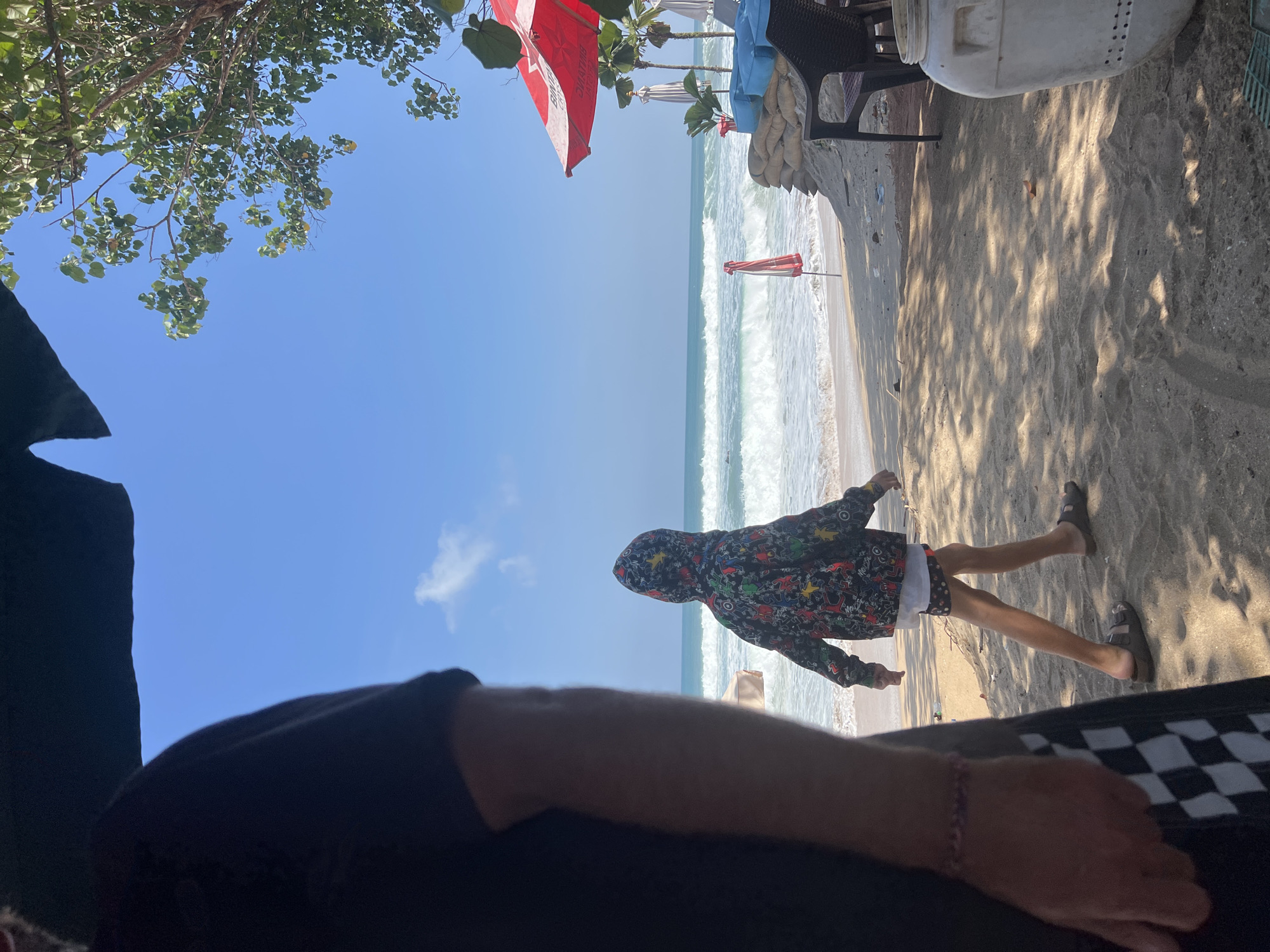 The image depicts a beach scene in Kuta, Indonesia. In the foreground, a person wearing a black t-shirt and checkered shorts is partially visible on the left, with their arm extended. To the right of the foreground figure, a child is walking away from the camera towards the ocean, wearing a hooded jacket with a colorful graphic print and patterned shorts. The child is wearing sandals. The beach itself is sandy, with some shadows cast by unseen objects. In the background, the ocean features moderate waves breaking on the shore. Along the shoreline, there are several beach umbrellas, one of which is a large red and white umbrella. Sandbags are stacked behind some seating areas, and a white plastic barrel is visible in the lower right corner of the image. The sky is clear and blue, with a few scattered clouds and a tree branch with green leaves extending into the frame from the upper right. The overall impression is a sunny day at a beach resort.