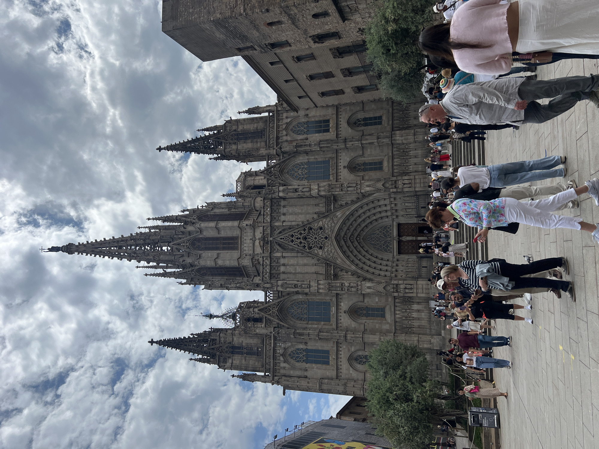 The image depicts the Gothic Cathedral of Barcelona, Spain, taken from street level, showing a crowd of tourists in front. The cathedral's facade is visible, including its pointed arches, spires, and ornate details. A stone building stands adjacent to the cathedral, partially obscuring the view of the sky, which features cloudy conditions. People are standing and walking on the stone paved street. A large group of people is waiting in line in front of the church. Trees are visible near the cathedral and crowd.