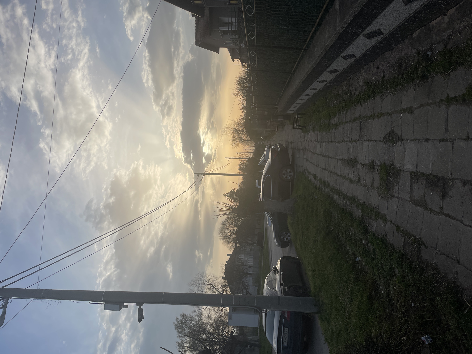 The image depicts a residential street in Aprilovo, Bulgaria, at sunset or sunrise. The sky is filled with clouds, with the sun's rays breaking through. A utility pole with wires runs along the left side of the frame. Parked cars line the right side of the street, which is paved with concrete blocks. A stone fence runs along the right side of the street, bordering a property with a house partially visible. Trees with bare branches are silhouetted against the bright sky.