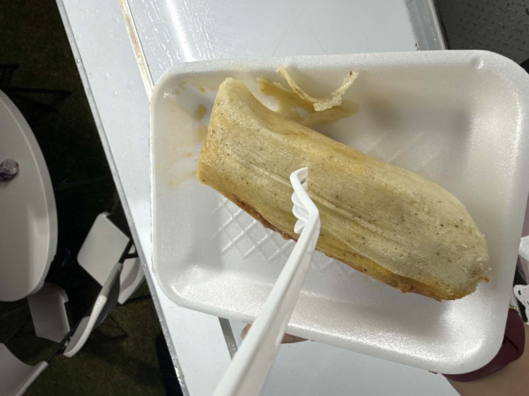 A close-up, high-angle shot reveals a tamale on a white polystyrene plate being held by someone with a white plastic utensil. The tamale is light brown with textured husks still partially encasing it. A few detached pieces of the husk sit near the top of the plate. A white table is visible in the background, along with some chairs and an area of grass. Based on the location provided, the scene is likely taking place in San Bernardino Tlaxcalancingo, Mexico.