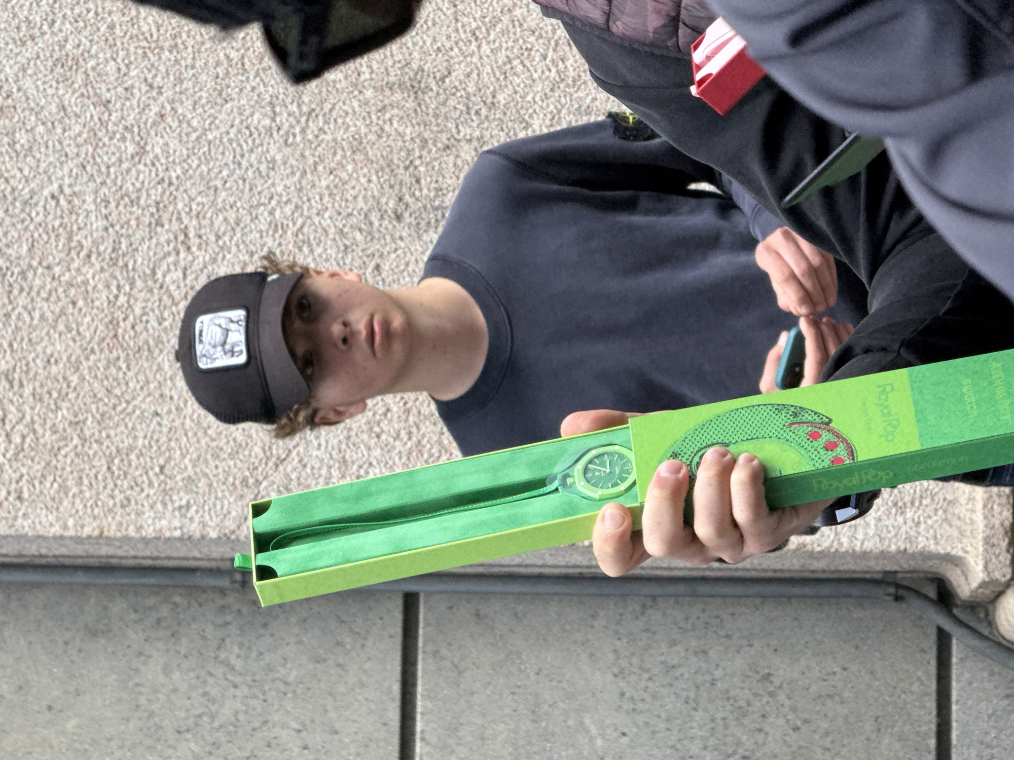 A medium, eye-level shot depicts two individuals and a green Royal Pop Swatch watch box. The setting appears to be outdoors, likely a city street in Milan, Italy.
A young man with light skin and short, curly blonde hair wearing a black baseball cap and a dark blue long-sleeved shirt stands to the left. He faces the camera directly, his expression neutral.
To his right, another person is partially visible, wearing a dark gray jacket and a red accessory pinned to the left side of the jacket. This person holds a green Royal Pop Swatch watch box in their right hand. The box is open, revealing a green wristwatch inside. The hand is fair-skinned with short fingernails. The product on the box appears to be RoyalPop and Swatch.
The background shows a beige concrete wall or ground. A dark gray ledge or curb is visible at the bottom, running horizontally across the frame.