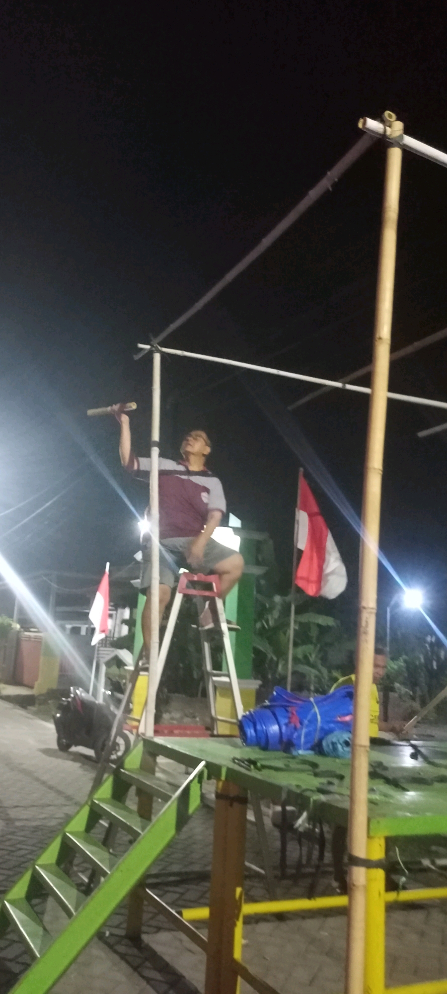 In Gempolklutuk, Indonesia, at night, a man stands on a ladder, holding a long, light-colored object, possibly a flag pole, and appears to be attaching it to a white PVC pipe structure that is supported by bamboo poles. He is wearing a maroon and gray t-shirt and gray shorts. To his right, a red and white flag is attached to a pole. In the foreground, a green table with a rolled-up blue tarp rests on it. A green staircase is visible to the left, and a motorcycle is parked nearby. Another person is partially visible to the far right, near a bright light source. The environment suggests an outdoor setting, possibly a temporary setup for an event.