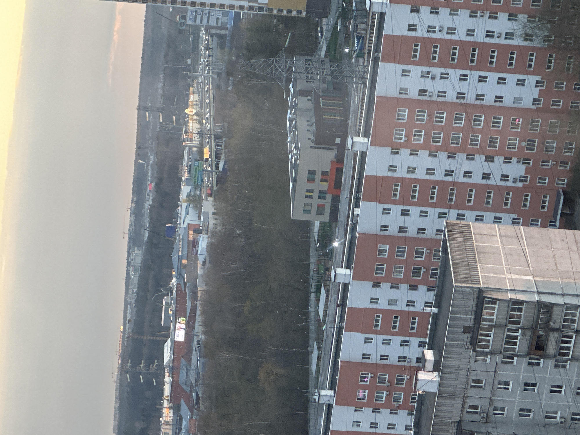 An aerial or high-angle view captures a cityscape in Otradnoye, Russia, under what appears to be an overcast sky or during dawn/dusk, with diffused light. The image is vertically oriented, giving the impression of a tilted horizon.
In the foreground and mid-ground on the right, several multi-story buildings dominate the frame. A prominent residential building features alternating horizontal bands of reddish-brown and light gray/white, with numerous uniformly sized windows and individual air conditioning units. One window in this building shows a distinct pink or magenta light. Adjacent to it, a darker, gray building with grid-like patterns and multiple windows is partially visible. Further back, another white building with colorful rectangular accents on its facade can be seen.
The middle ground is characterized by a significant expanse of dense, dark-foliaged trees, likely a park or wooded area, separating the foreground buildings from the distant horizon. Overhead power lines and a large metallic electricity pylon cut across this wooded area.
Along the edges of the wooded area and in the background, more urban development is visible, including roads, streetlights, and smaller buildings. Hints of distant construction cranes can be seen against the horizon.
Visible text includes a green sign that appears to say "Сбербанк" (Sberbank) and a yellow sign with "Рынок Строительный" (Stroitelny Market - Construction Market), both indicating commercial establishments. No specific human activity is discernible due to the high vantage point. The scene depicts a typical urban landscape with residential and commercial structures integrated with natural green spaces.