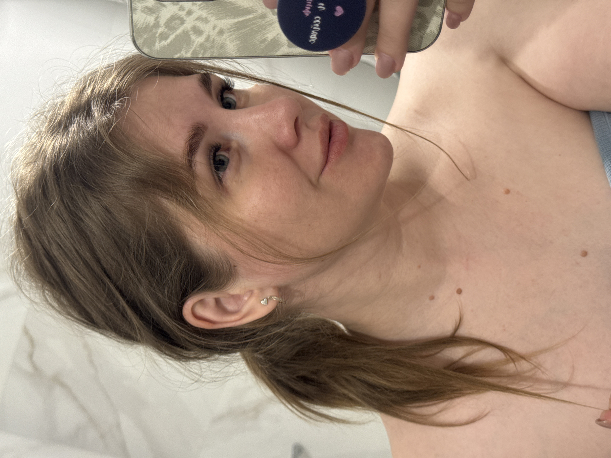 A woman is taking a selfie. She has light brown hair, blue eyes, and is wearing earrings. She is holding a phone with a patterned case and a blue pop socket. The background appears to be a white wall. The location is Ферма, Russia.