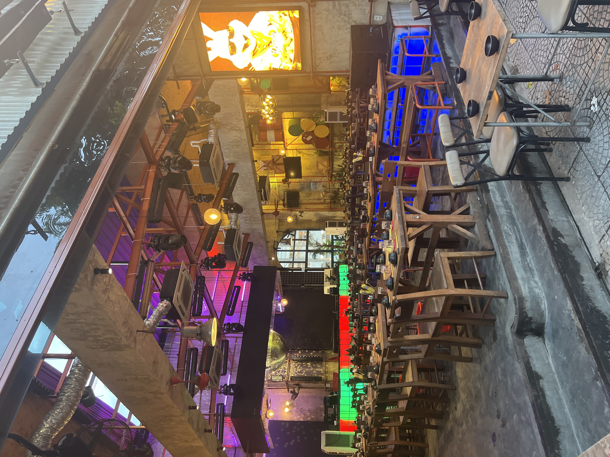 The image depicts the interior of a restaurant or bar in Thuận Hóa Ward, Vietnam, with an industrial-style design. The space features rows of wooden tables and chairs set up for dining. Above the dining area, a complex ceiling structure is visible, with exposed ductwork, lighting fixtures, and what appears to be a large screen or stage area in the background. The lighting is a mix of ambient and accent lighting, with some areas illuminated by purple and pink lights, and others by warm yellow tones. A large LED screen displays an animated character of a rabbit wearing sunglasses. The overall atmosphere suggests a lively entertainment venue, possibly a karaoke bar or a restaurant with a performance space. The flooring is a mix of concrete and tiled surfaces. There are no people visible in the scene.