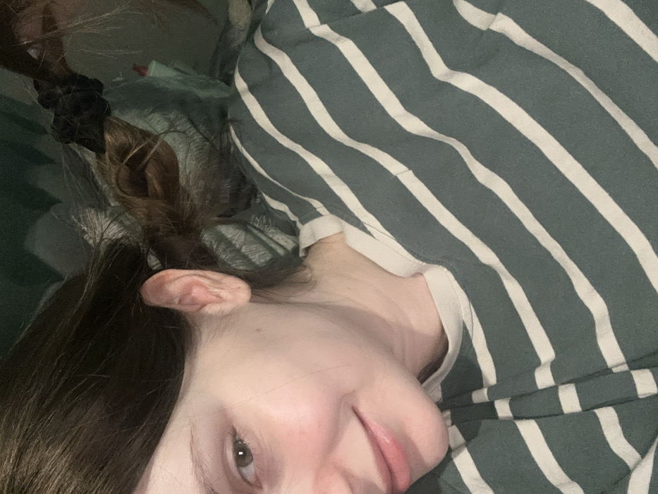 A person lies down in a striped shirt. Their hair is brunette, with a scrunchie holding some of it up. One ear is visible. The person has fair skin, a slight smile, and light eye color. The bedding or clothing below the person appears to be green. The image is taken from a slightly overhead, angled perspective.