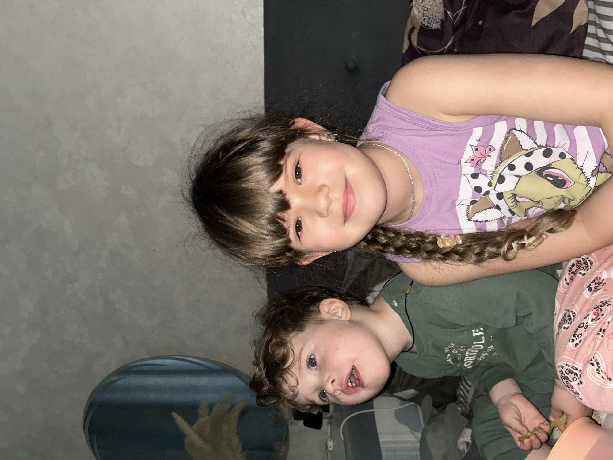 Two young children are lying down. The elder child, a girl, has brown hair with bangs and a braid. She is wearing a light purple tank top with an illustration of a cat and butterflies. The younger child, a boy, has curly brown hair and wears a green long-sleeved shirt. He appears to be holding a small object in his hand. A fishbowl is visible in the lower left corner of the image. The background includes a gray wall and a dark gray upholstered headboard. The location, Klin, Russia, is not discernible from the image.