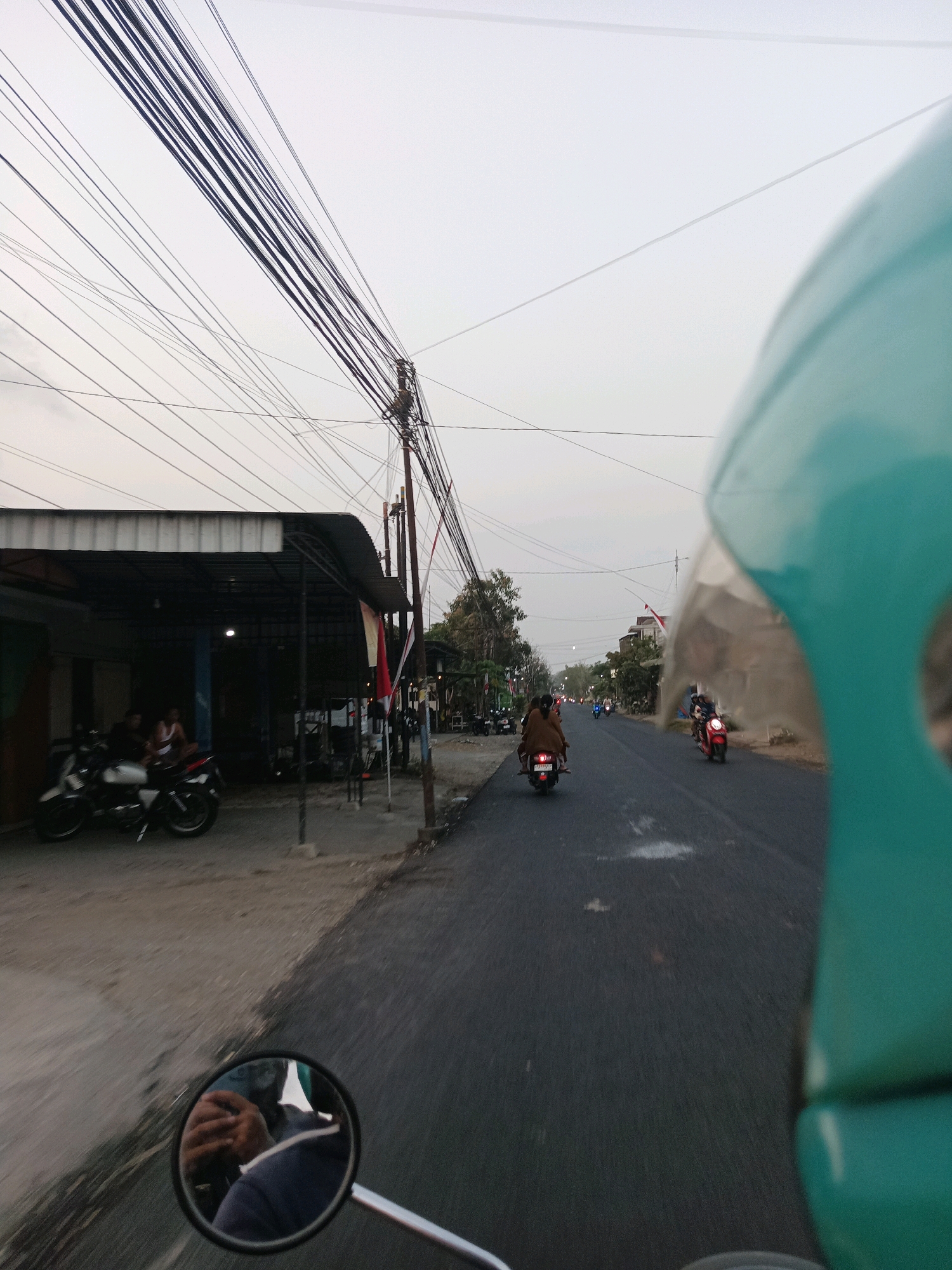 The image depicts a street scene in Gedangan, Indonesia, as viewed from the perspective of someone riding a motorcycle. In the foreground, a portion of a teal-colored helmet is visible on the right side. A motorcycle's side mirror is centrally positioned, reflecting the rider's hands and forearms, which are clasped together. The road surface appears wet in places.
To the left of the road, a covered structure, possibly a shop or workshop, is situated. Several motorcycles are parked under this shelter, and two individuals are seated there. In front of the structure, more motorcycles are parked on the ground. Overhead, a dense network of electrical wires crisscrosses the sky, supported by utility poles.
Further down the road, other motorcycles are visible, with riders on them. One motorcycle ahead and to the left has two occupants, both wearing brown clothing. Another motorcycle, further ahead and to the right, is red. Trees line the right side of the road in the distance. The sky is overcast, suggesting either early morning or late afternoon. The overall impression is of a typical roadside scene in a bustling Indonesian town or city.